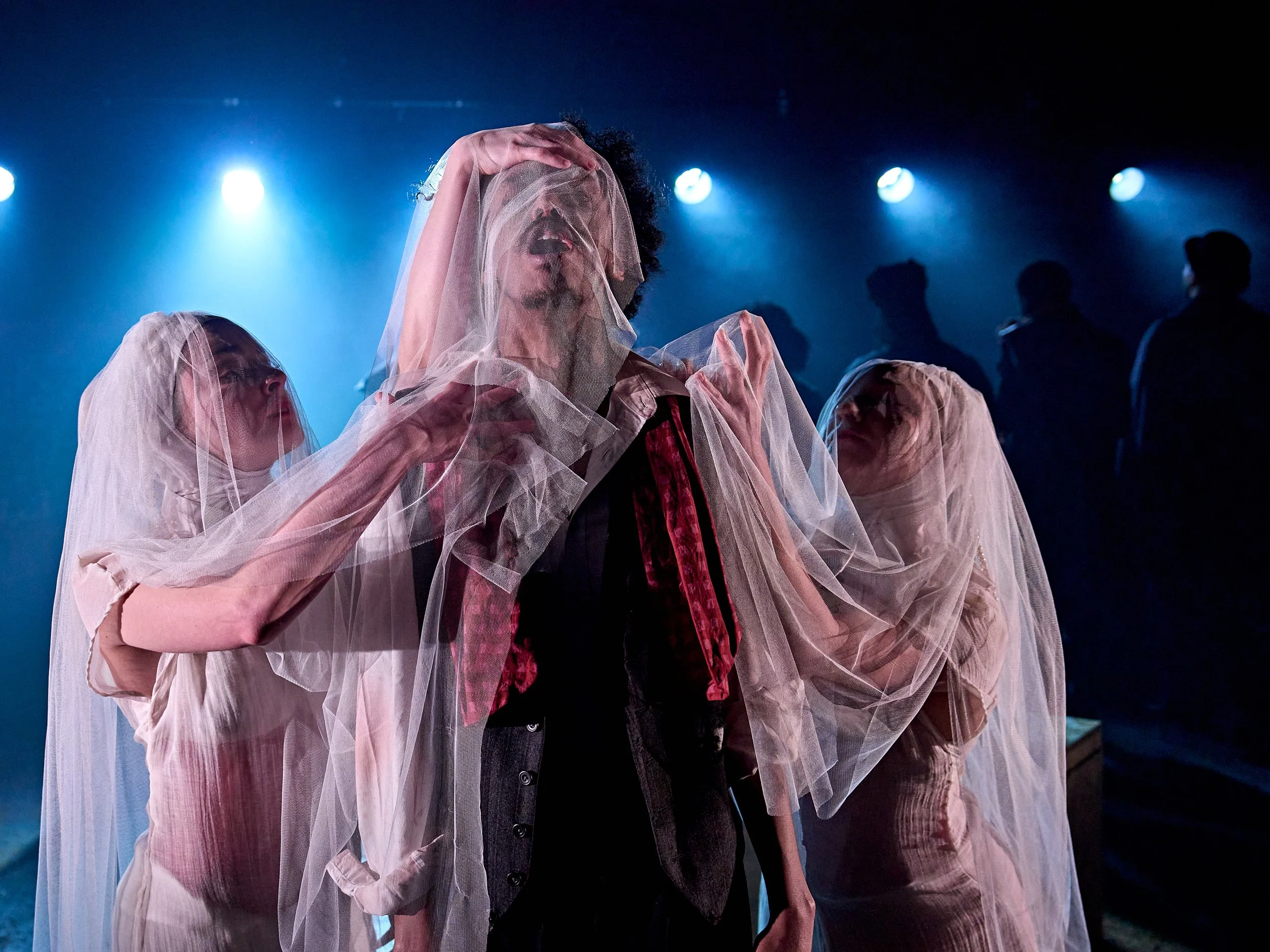 Person with curly hair wearing a pink sheer fabric veil supporting their head and face, in a dark setting with blue stage lights and silhouettes of people in the background.