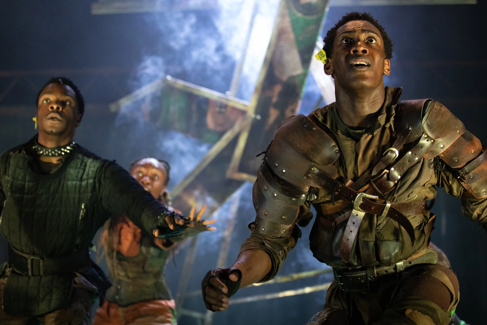 A group of people dressed in rugged, futuristic costumes with armor and tactical gear, looking upward with expressions of concern or awe inside a dimly lit, industrial setting with smoke and metal structures.