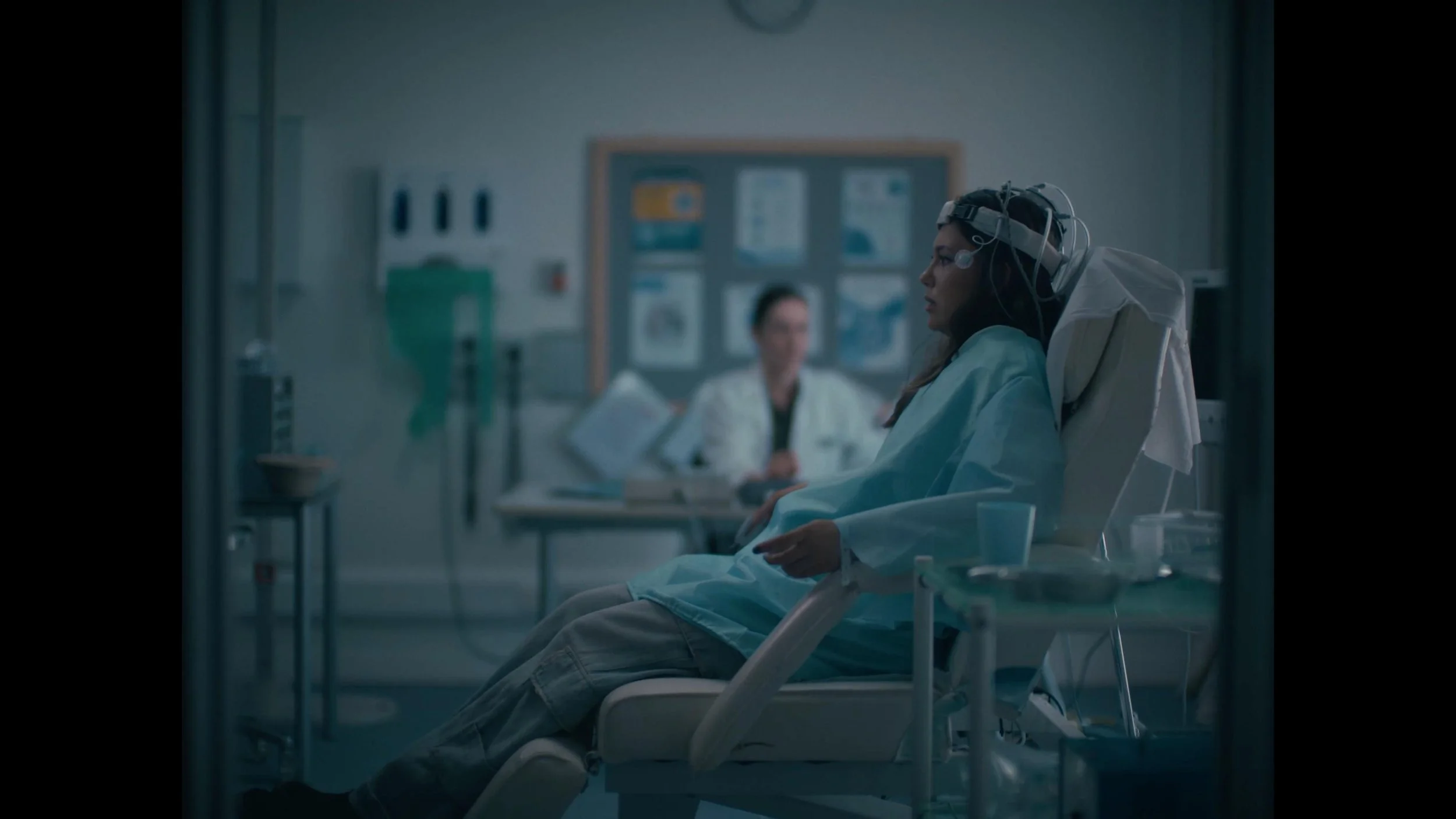 A woman in a hospital bed with medical equipment on her head, sitting in a hospital room. A doctor or nurse is blurred in the background, sitting at a desk.