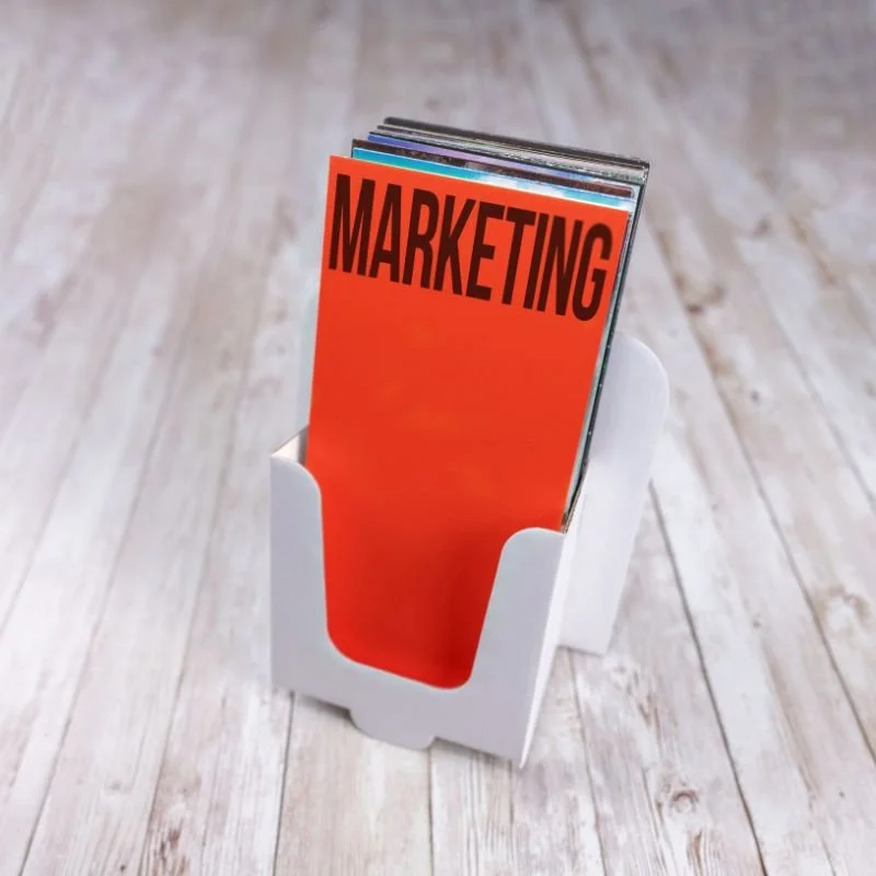 Brochure or magazine with the title "MARKETING" in bold black letters on a red background, held in a white cardholder on a wooden surface.
