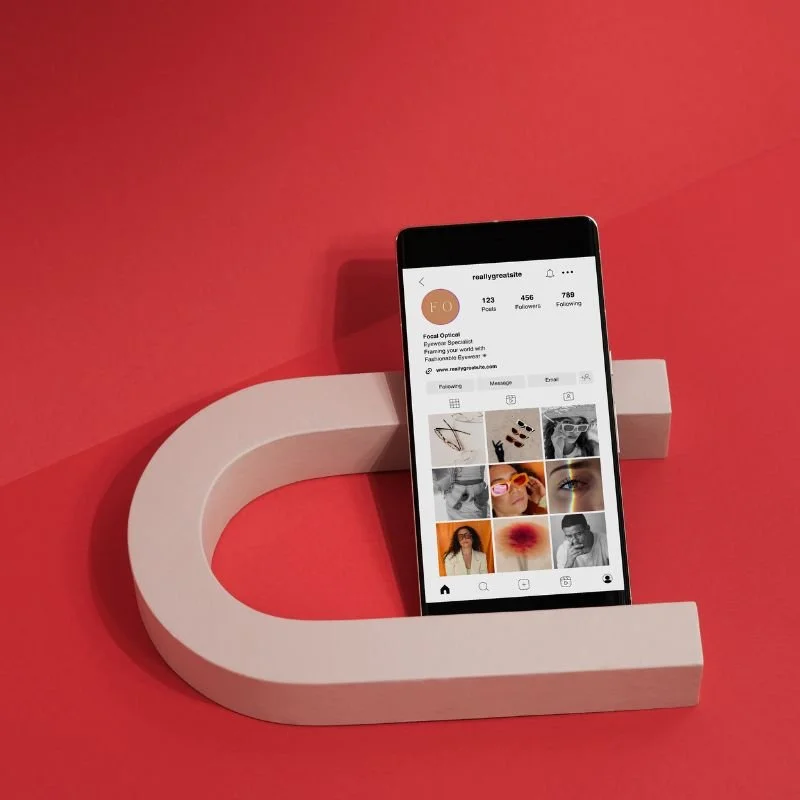 Smartphone displaying an Instagram profile, placed on a pink surface with a white letter C-shaped object.