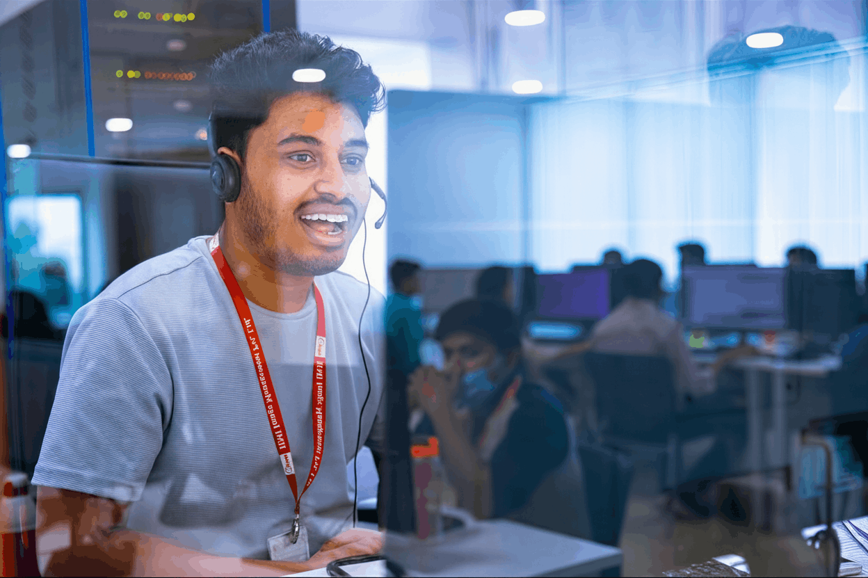 A young man with dark hair wearing a headset and a white shirt, smiling while working at a computer in a modern office environment with other people working in the background.