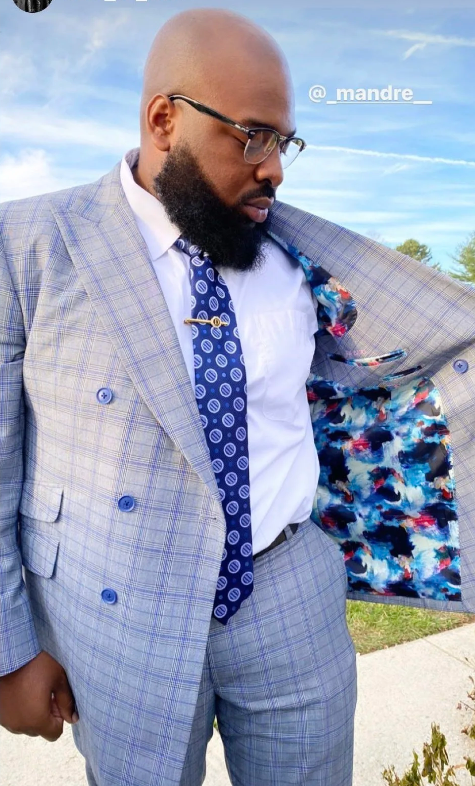 Man in a gray plaid suit with a colorful floral blazer lining, white shirt, navy blue patterned tie, glasses, and a beard, standing outdoors against a blue sky background.