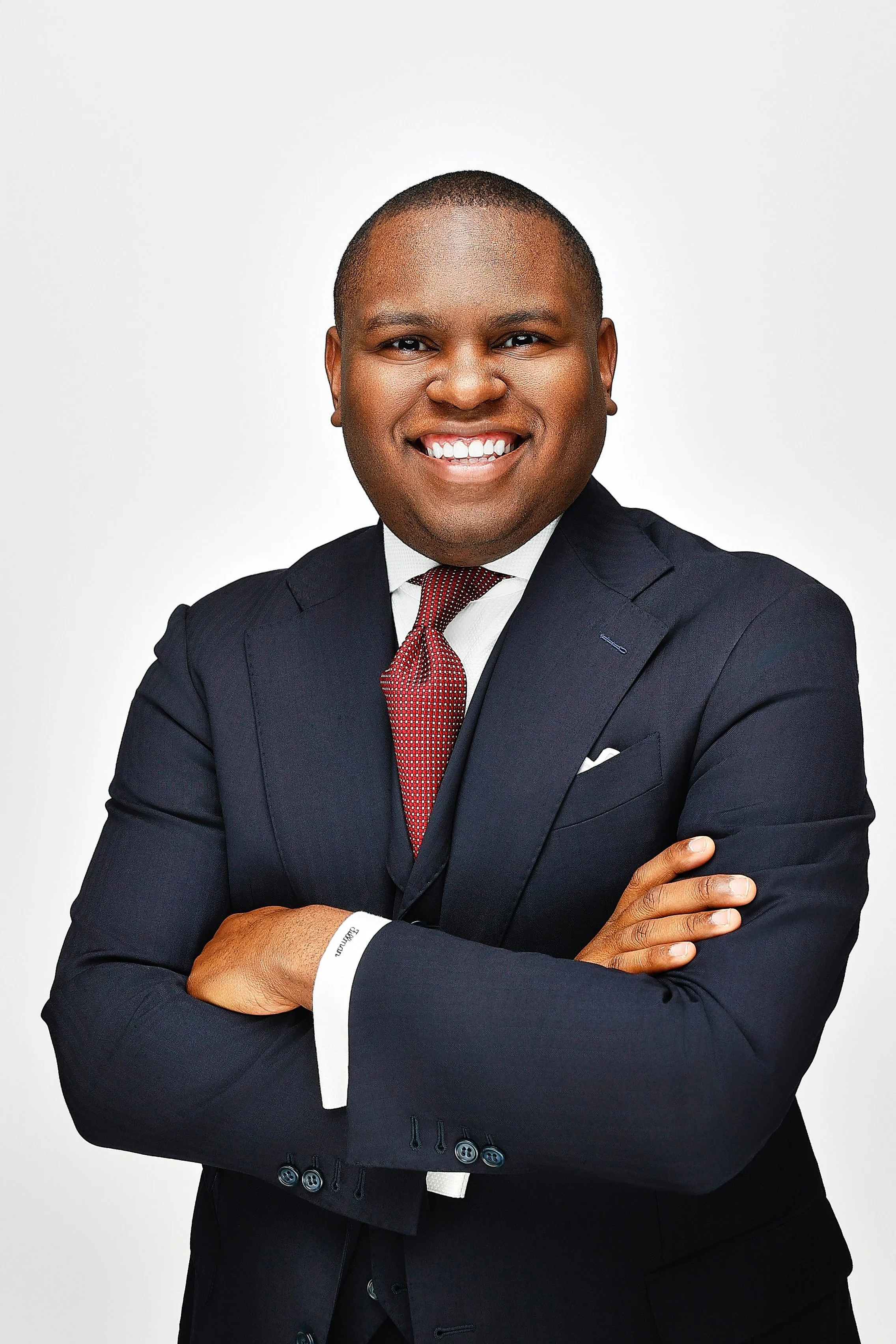 Professional man wearing a navy suit with a white shirt and a red tie, arms crossed, smiling confidently against a plain white background.