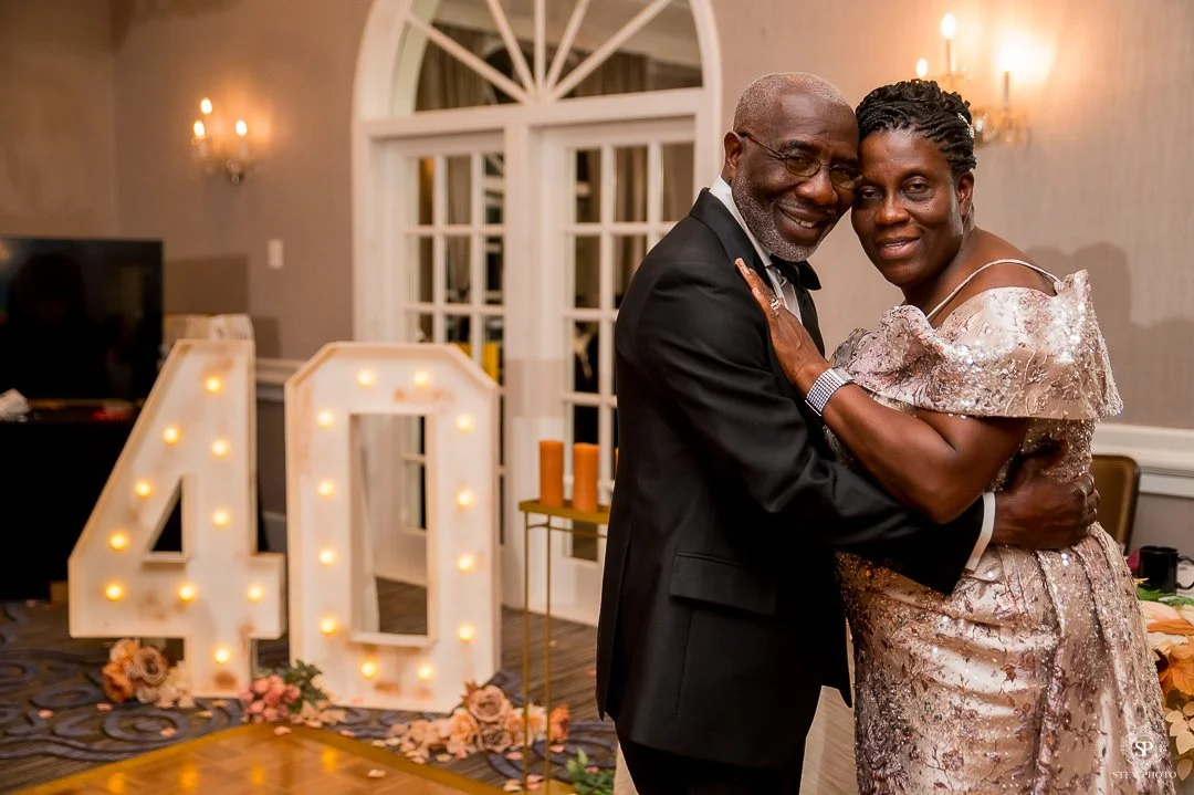 A happy couple dancing and hugging at a celebration, with large illuminated number 40 and floral decorations in the background.
