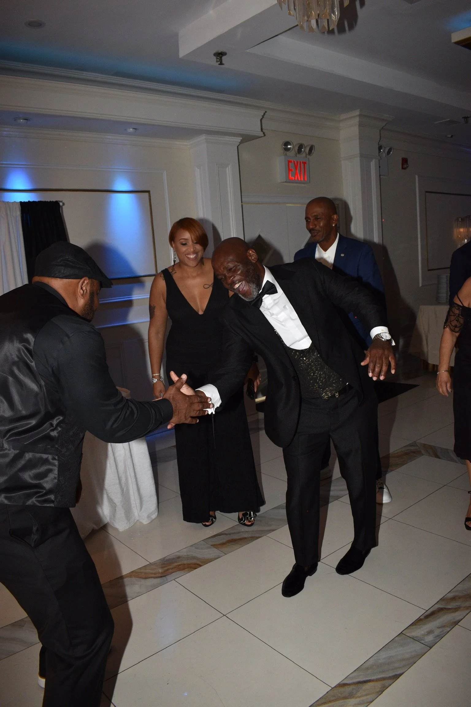 A group of people dancing and socializing at an indoor event, with a man in a tuxedo and a woman in a black dress prominent in the foreground, smiling and having a good time.