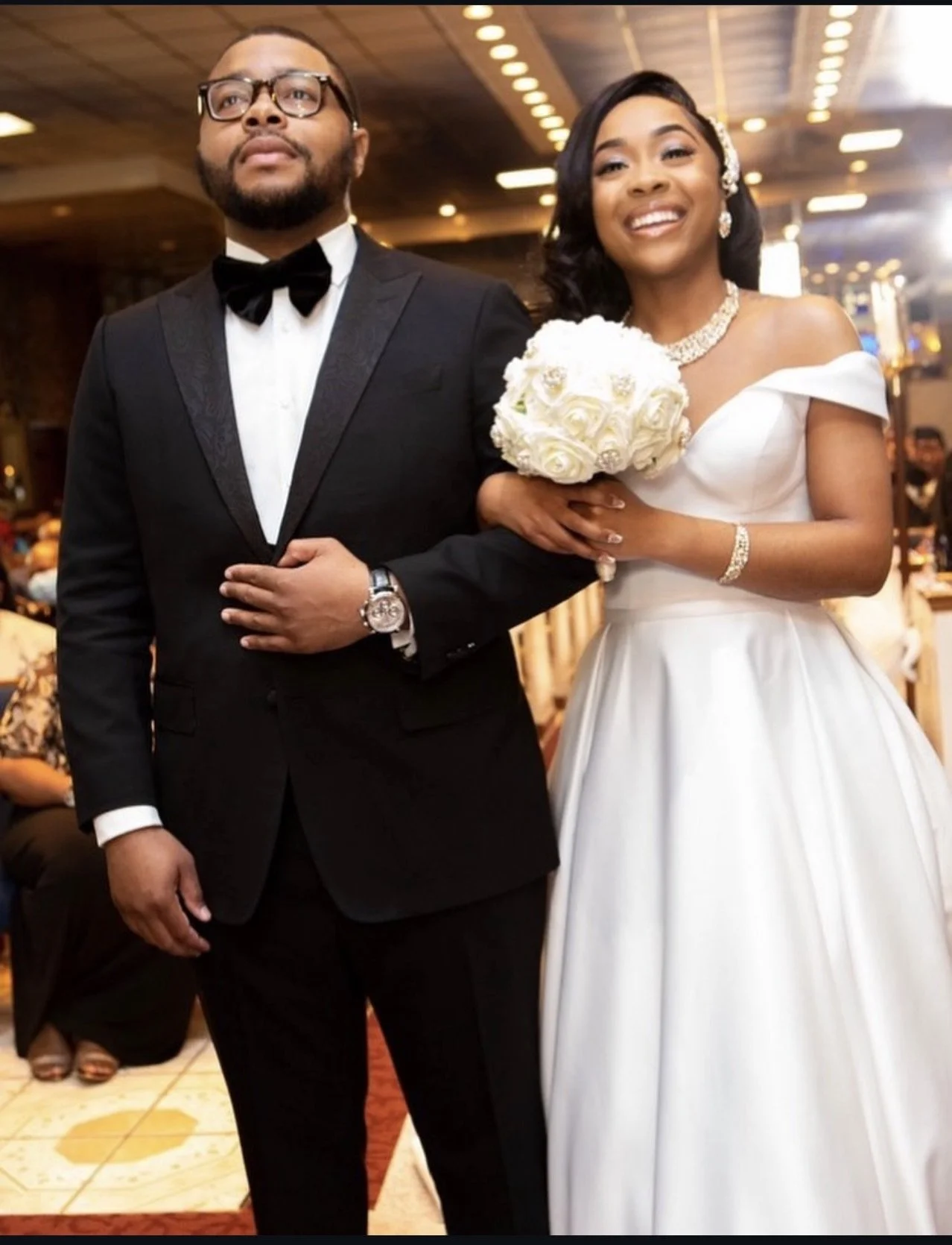 A bride and groom standing together at a wedding reception. The groom is wearing a black tuxedo and glasses, and the bride is in a white wedding gown holding a bouquet of white roses, smiling happily.