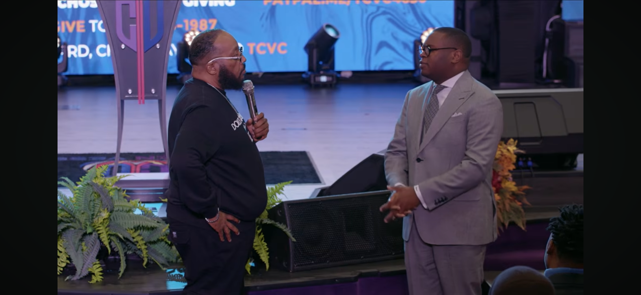 Two men are standing and facing each other in a church or conference setting. One man is holding a microphone and wearing a black shirt. The other man is dressed in a gray suit and glasses. There are plants and a large screen or backdrop behind them.