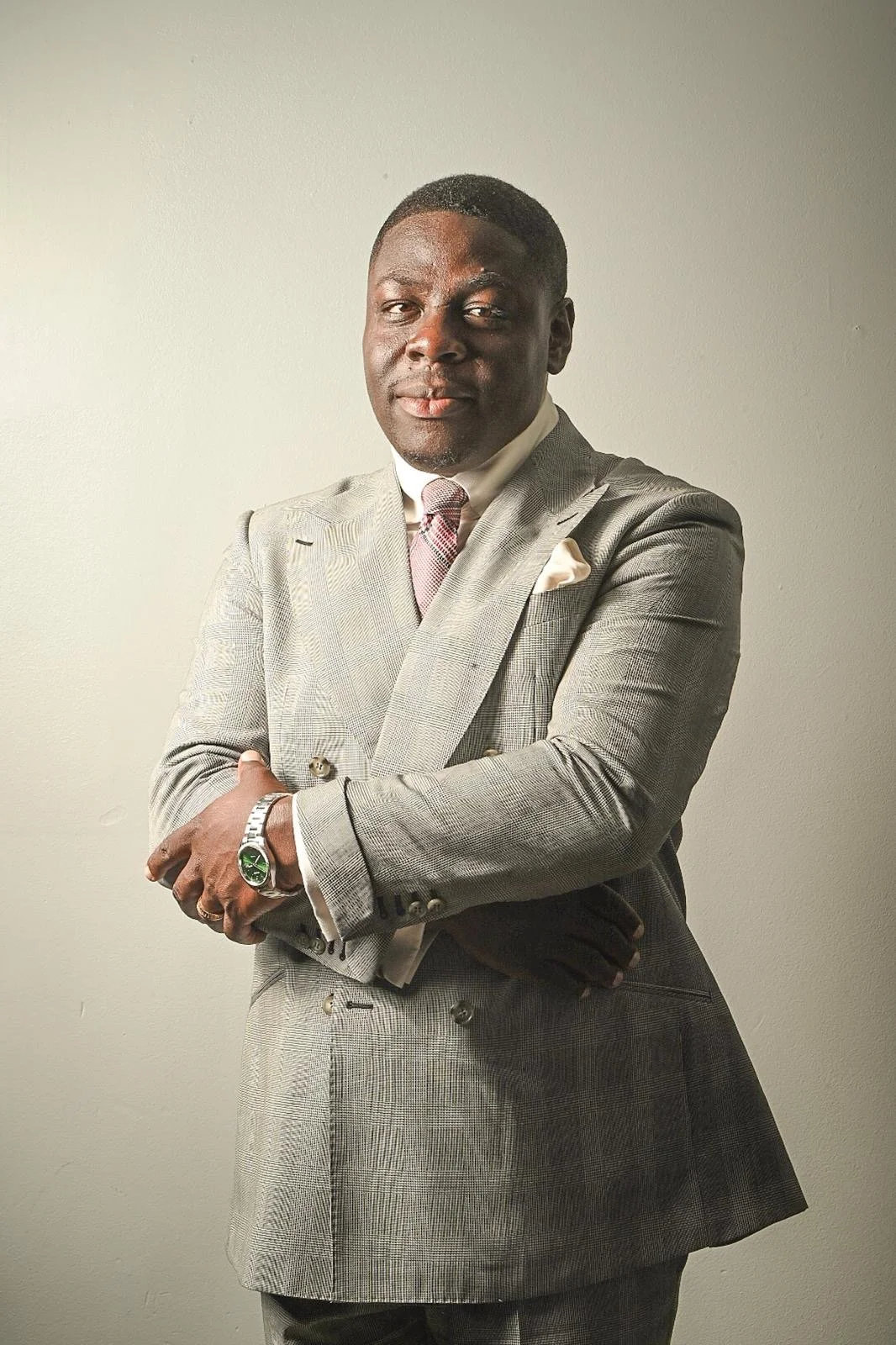 A man in a light gray plaid suit with a pink tie and white shirt, standing with arms crossed in front of a plain white wall.