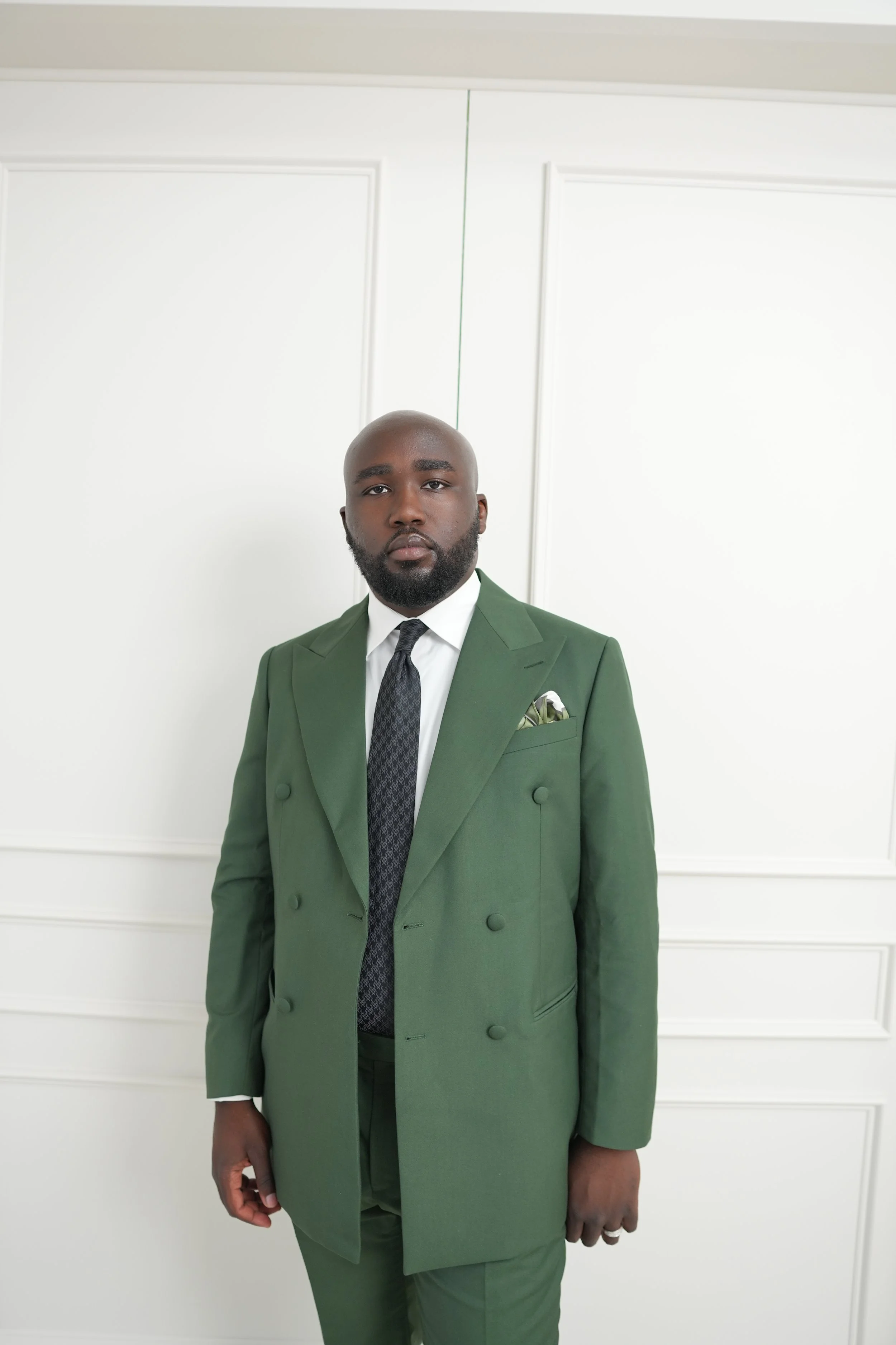 A man wearing a green suit with a white shirt, black tie, and a pocket square stands in front of a plain white wall with paneling. He has a shaved head and a beard, looking directly at the camera.