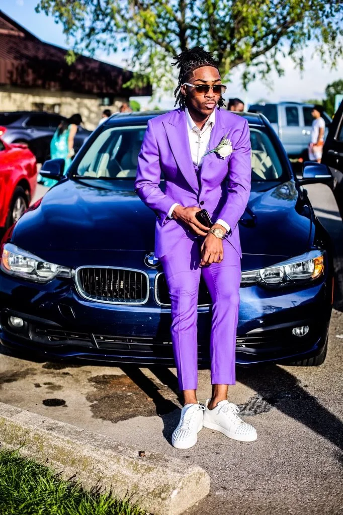 Man dressed in a purple suit and white sneakers leaning against a black BMW in a parking lot, holding a phone, wearing sunglasses, with other cars and people in the background on a sunny day.