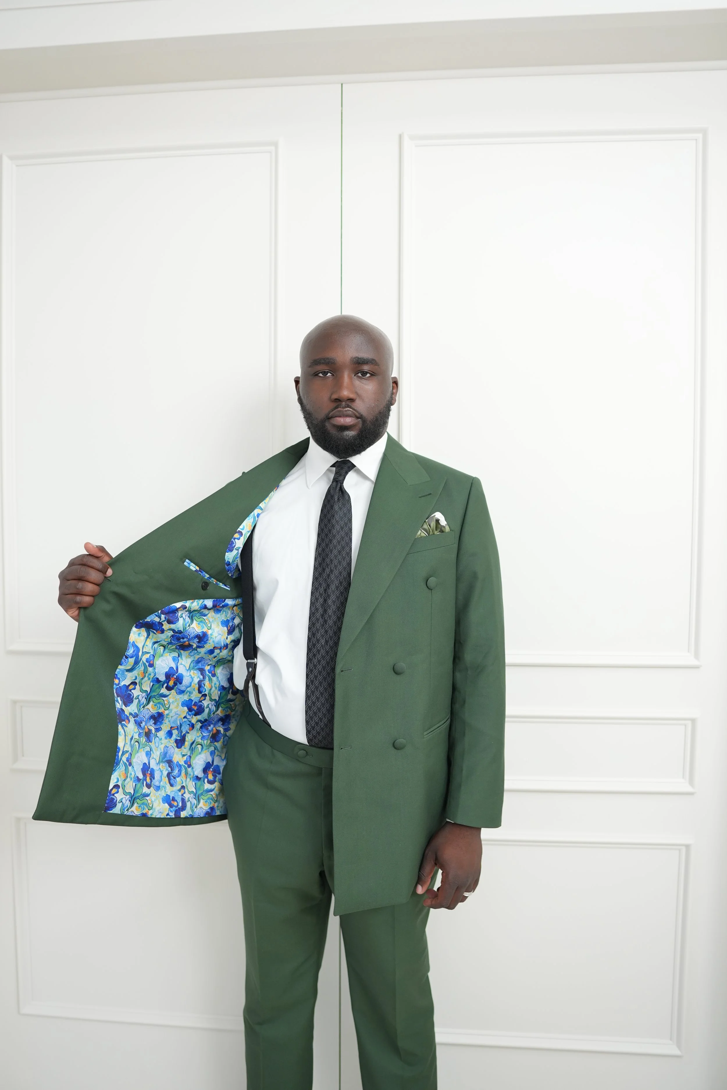 A man in a green suit holding open the inner lining of his jacket, which has a colorful floral pattern, against a white paneled wall.