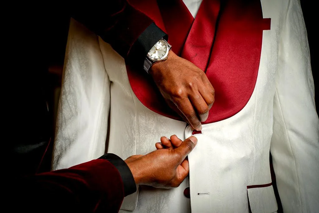 A person in a white tuxedo with black and red accents is being assisted with buttoning the suit by another person. The individual in the tuxedo is wearing a watch with a silver band.