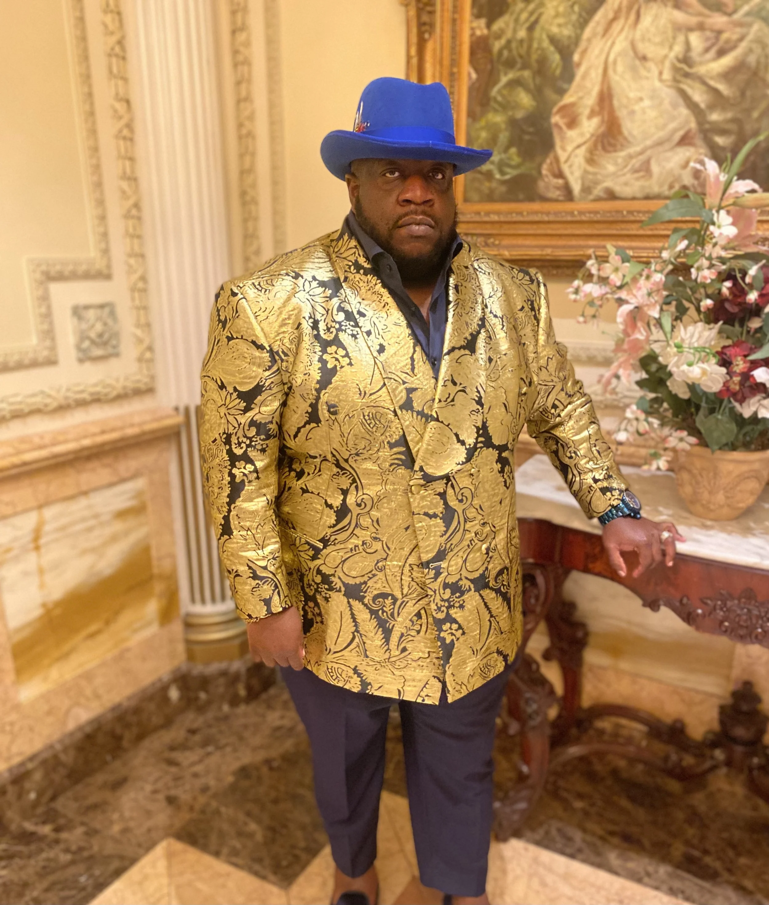 A man wearing a bright blue hat with a feather and a shiny gold patterned blazer. He is standing in an ornate, decorated room with floral arrangements and a large painting in the background.