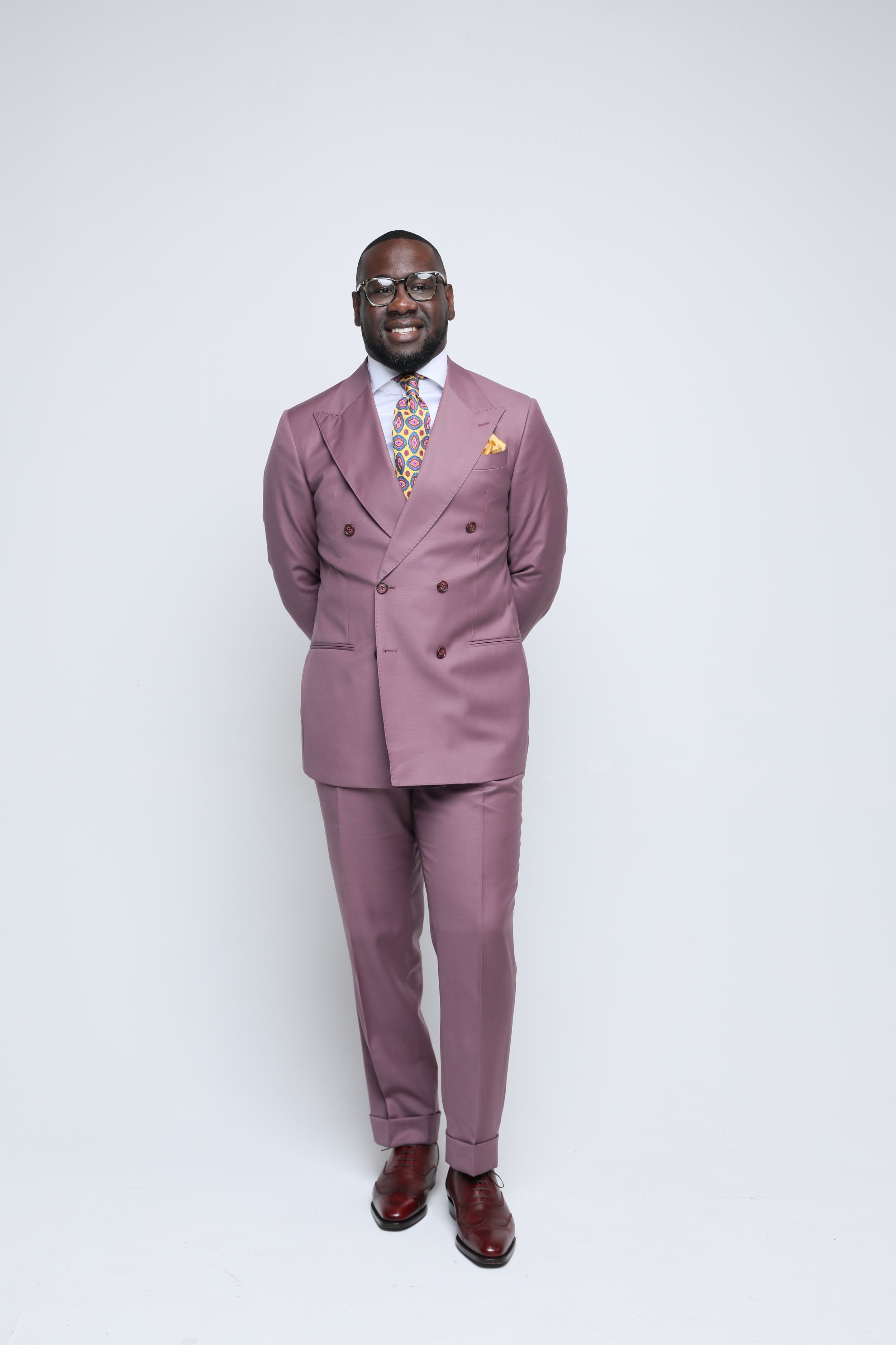 A man wearing a pink double-breasted suit with matching pants, a colorful tie, a pocket square, and brown shoes is standing in front of a plain white background.