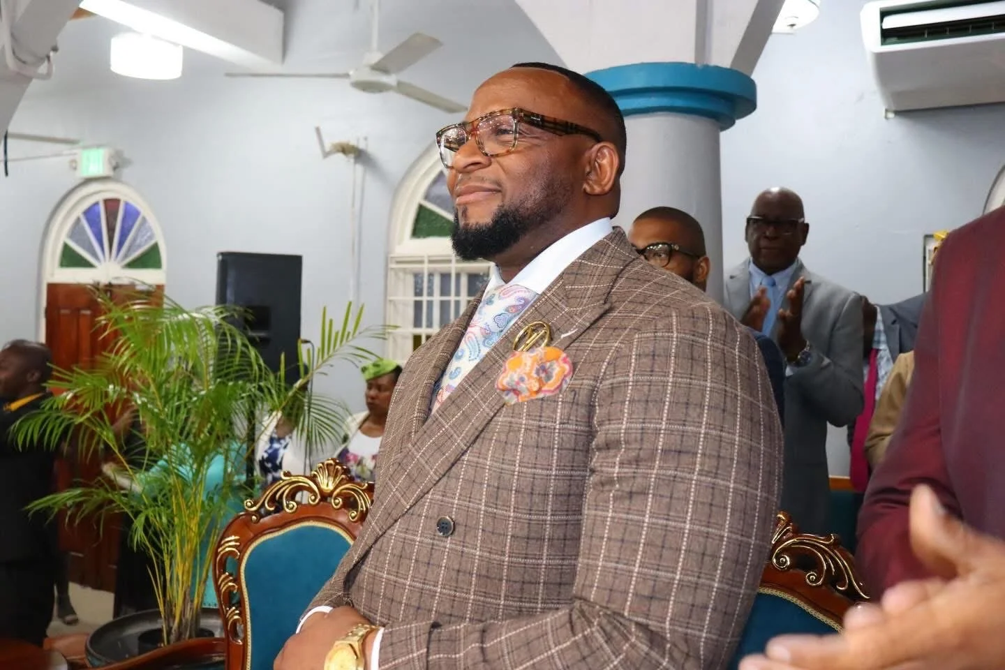 A man in a brown plaid suit with glasses, standing with a slight smile, in a room with people clapping, plants, and decorative chairs.