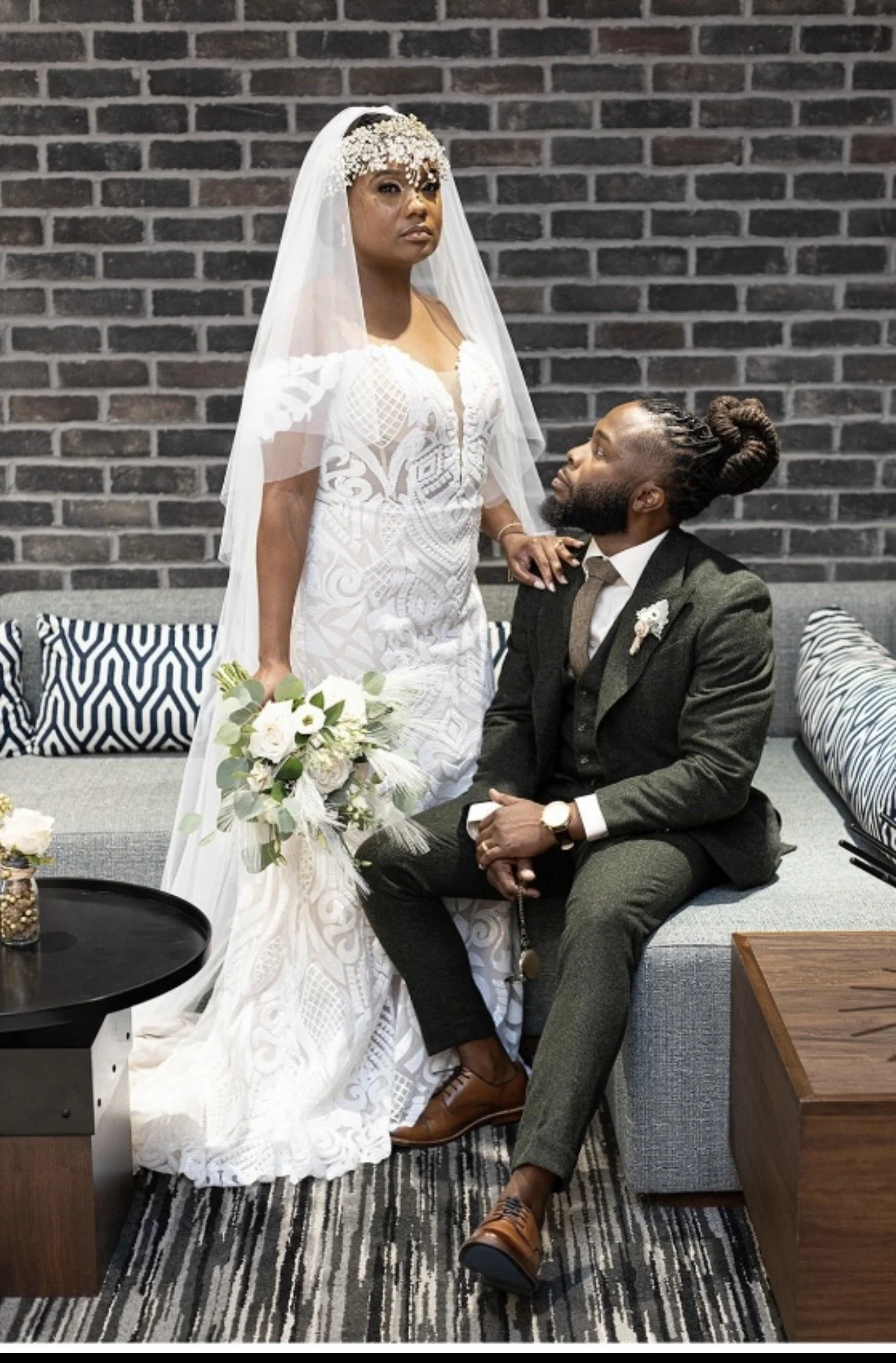 A bride in a white lace wedding dress with a veil and floral headpiece stands next to a seated groom in a dark gray suit with a white shirt and tie, inside a modern room with a dark brick wall background and striped rug.