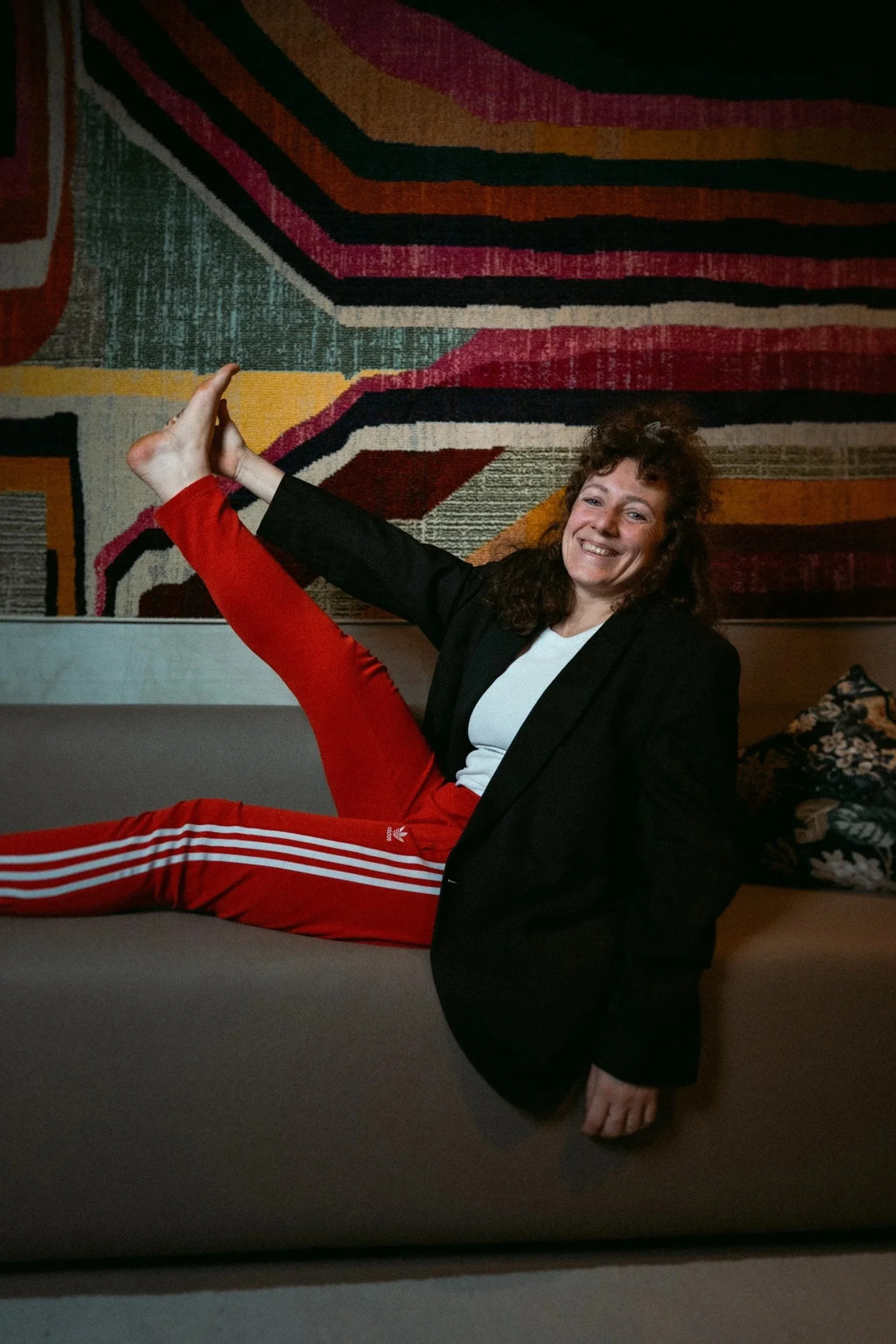 Coco von high on prana sitzt lachend in einem schwarzen Blazer und roter Sporthose sitzt auf dem Boden vor einem bunten, geometrischen Wandteppich.