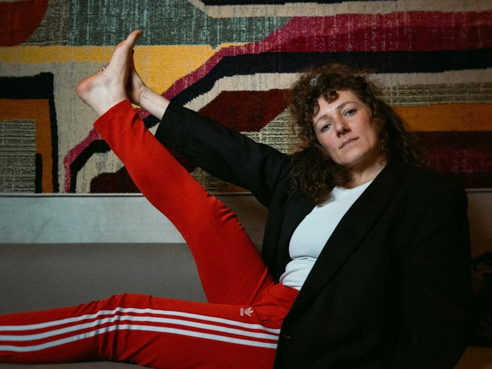 Coco von high on prana sitzt auf einer Couch vor buntem Hintergrund und hält ein Bein nach oben