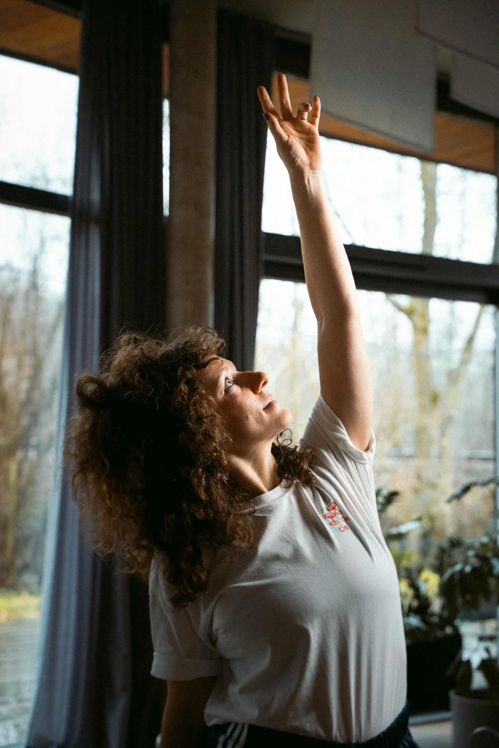 Eine Frau mit hellbraunen, lockigen Haaren hebt während einer Yogaasana ihre Hand in die Luft, während sie nach oben schaut. Sie befindet sich im Inneren eines Raumes mit großen Fenstern, durch die Bäume im Hintergrund sichtbar sind.