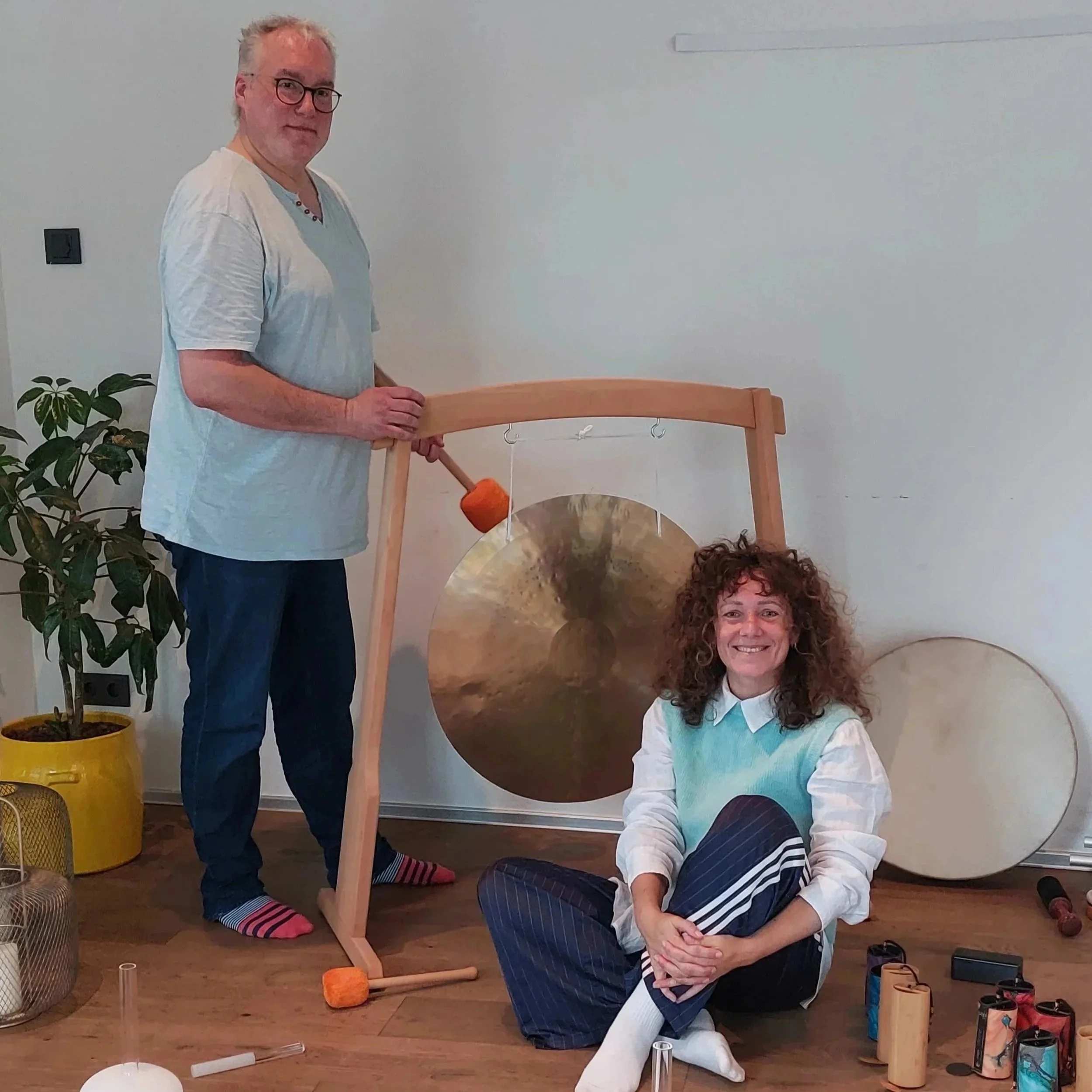 Zwei Personen im Raum mit einem großen Gong und anderen Musikinstrumenten, bei einer musikalischen oder kreativen Aktivität, die auf einer weißen Wand im Hintergrund dargestellt wird.