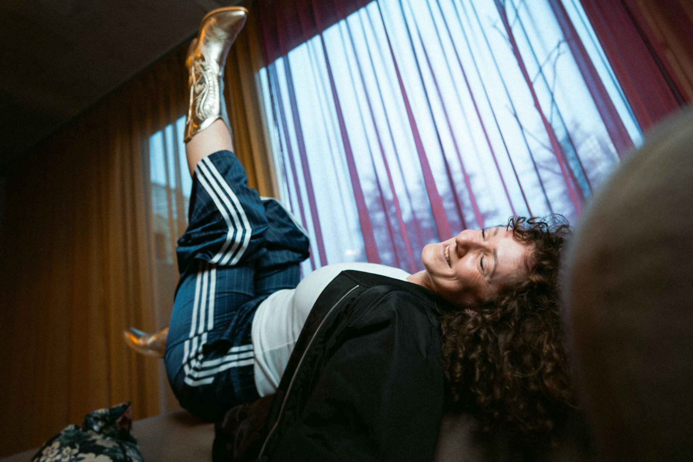 Coco von high on prana liegt lachend auf einem Sofa in einem Raum mit großen Fenstern und roten Vorhängen. Sie trägt eine schwarze Jacke, ein weißes Shirt und schwarze Adidas-Hose.
