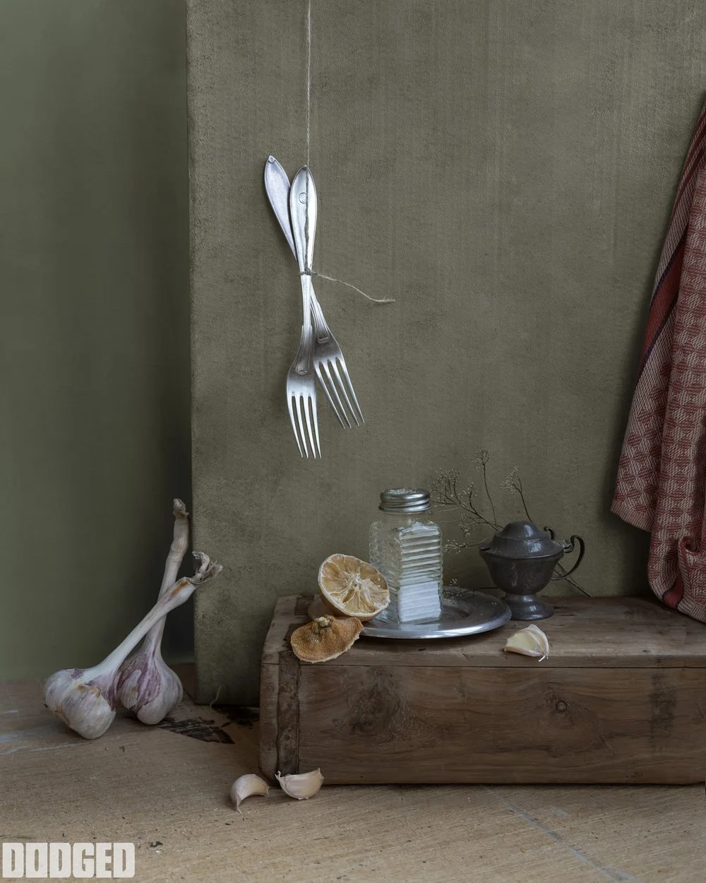 Two garlic bulbs on the floor, a wooden block with garlic and lemon slices, a glass jar, a metal sugar bowl, and other small objects on top of the wooden block, with two silver forks hanging on string against a green wall.