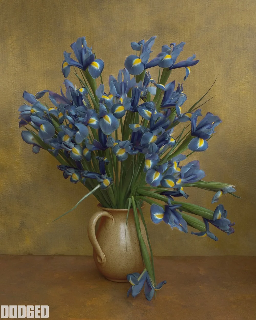A beige vase filled with a large bouquet of blue and yellow iris flowers placed on a brown surface against a muted yellow background.