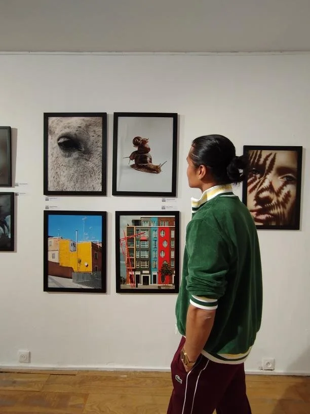 A person standing in an art gallery looking at framed photographs and paintings on a white wall. The person is wearing a green jacket and maroon pants.