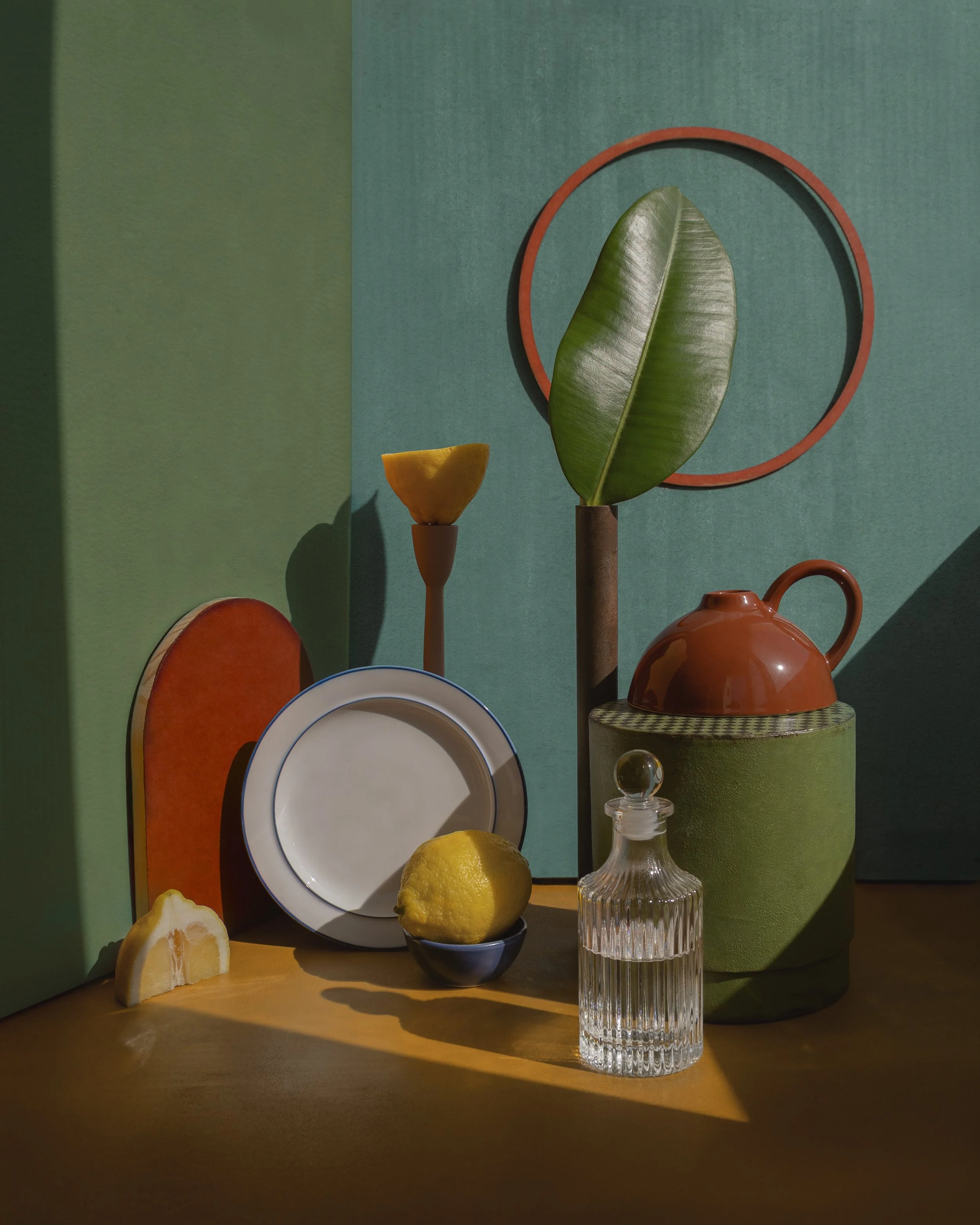 A still life arrangement with lemon slices, a whole lemon, a white plate with a blue rim, a small glass jar, a large green vase, and decorative objects on a wooden surface against a green and blue background.