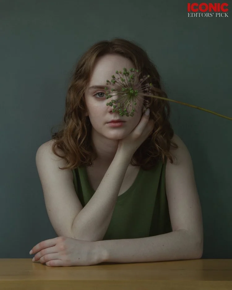 A young woman with curly red hair wearing a green sleeveless top, resting her chin on her hand, with a large flower covering one eye, sitting at a wooden table against a plain gray background.