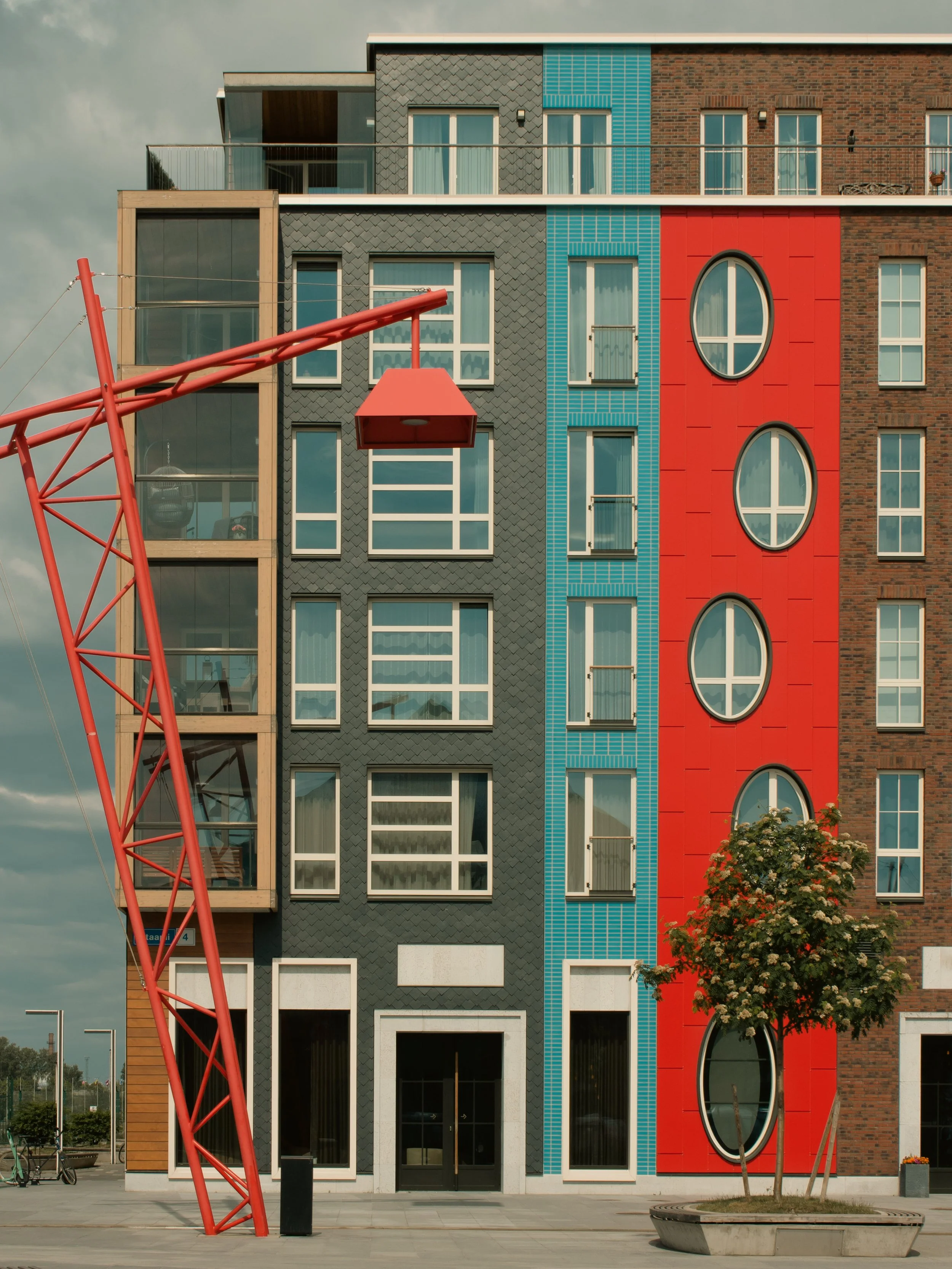 Multicolored modern apartment building with black, red, blue, and brick sections, featuring round and rectangular windows, with red sculpture in foreground and trees nearby.