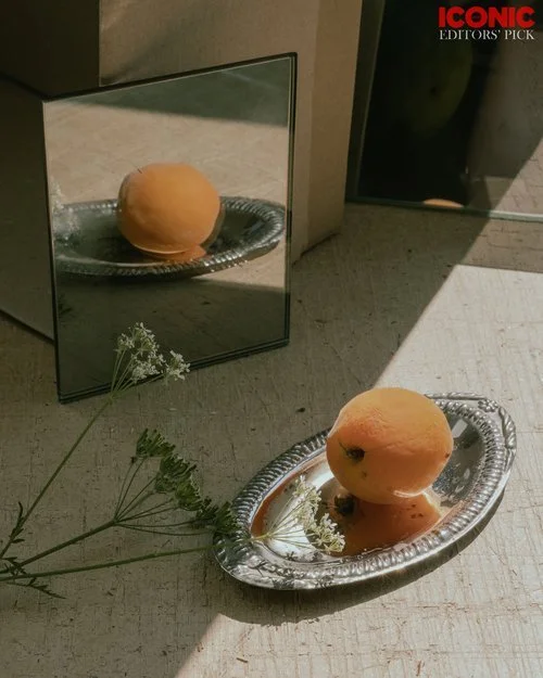 A peach placed on a silver tray on the floor, with a mirror behind reflecting the peach. There are some white flowers in the foreground, and a shadow cast on the wall and floor.