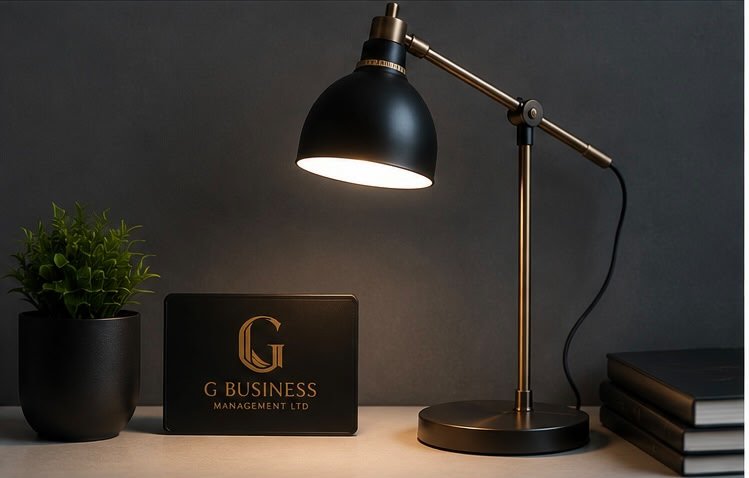 Desk with a black desk lamp, a small green plant in a black pot, a black sign with a gold 'G' and text reading 'G Business Management Ltd.', and two stacked black notebooks or folders.