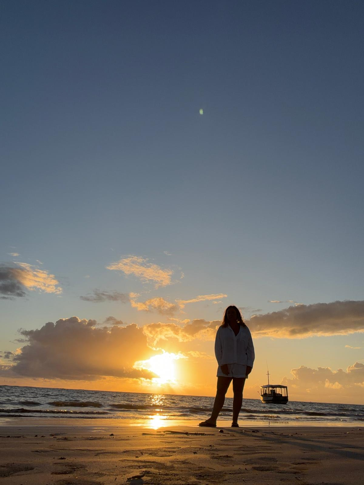 A person standing barefoot on a beach during sunset, with the sun near the horizon, clouds in the sky, a boat in the water, and the person silhouetted against the sunset.
