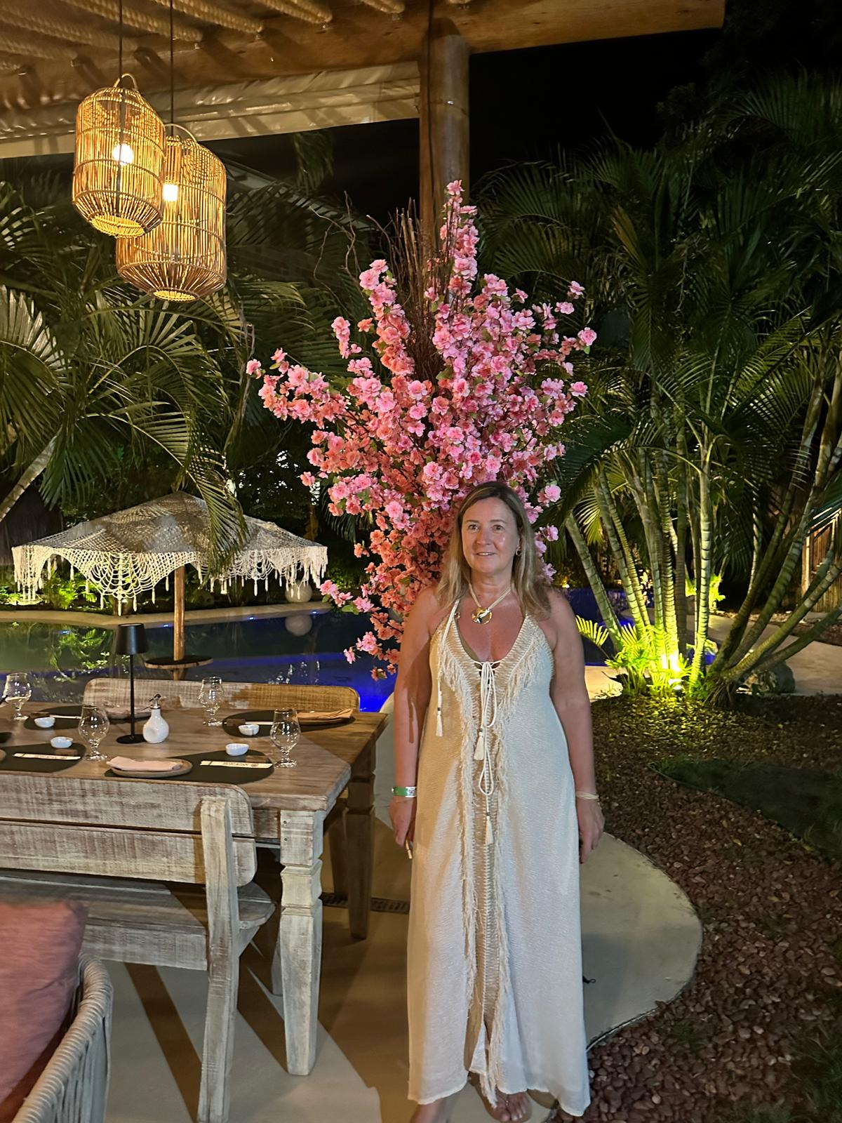 A woman standing in front of a pink cherry blossom tree at an outdoor dining area at night. The scene includes a wooden table set for dining with glasses, plates, and napkins, and decorative lighting such as hanging lanterns and a table lamp. There is lush greenery and a pool in the background.
