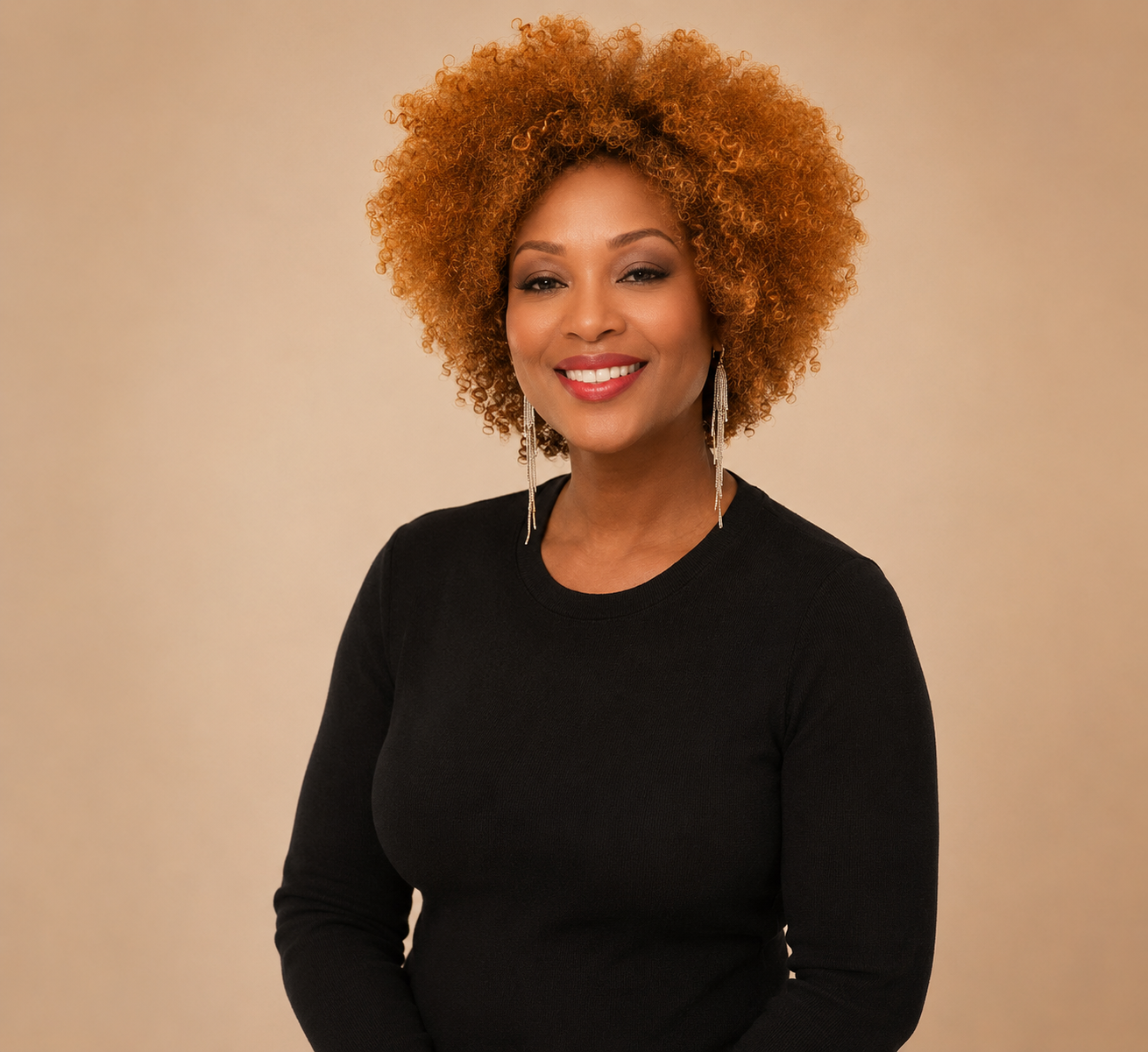 Portrait of a smiling woman with curly red hair, wearing a black top and long silver earrings, standing against a beige background.