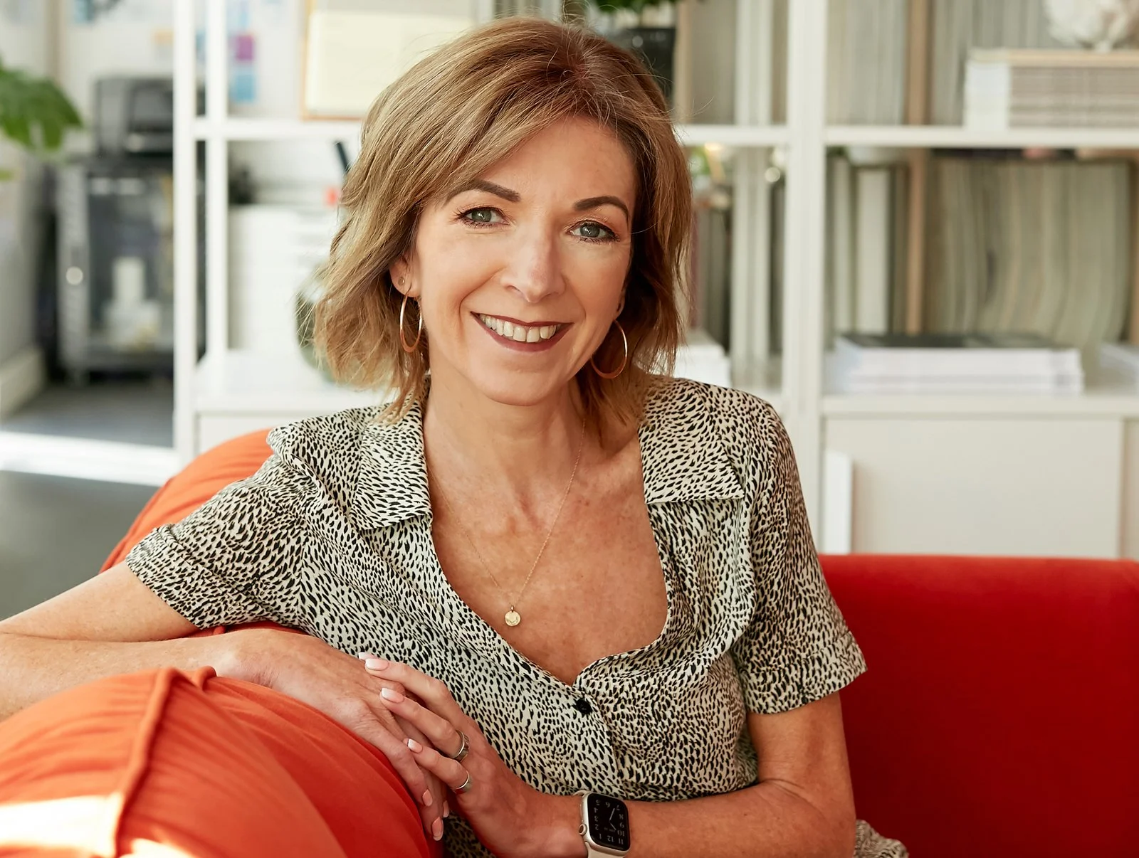 Nik Dillon. Smiling woman with short, wavy hair, wearing a leopard print shirt, jewellery, and a watch, sitting on a red couch in a bright room with shelves of books and a plant in the background.