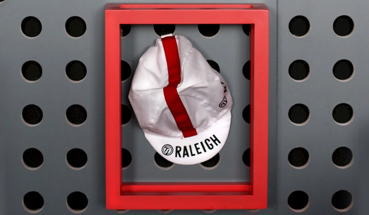 A white cap with red stripe and the word "Raleigh" hangs on a red framed hook on a gray pegboard.