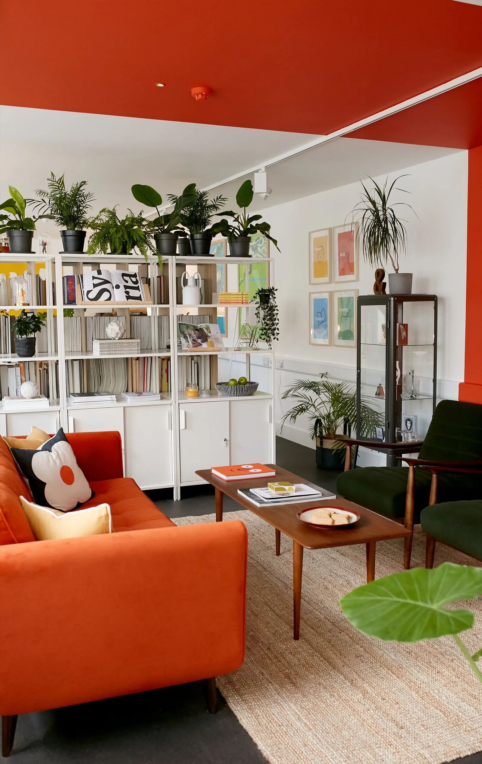 Alkamee Brand and Design Studio. A cosy meeting room with an orange sofa, green armchair, wooden coffee table, bookshelf with plants, and framed colourful art on the white walls.