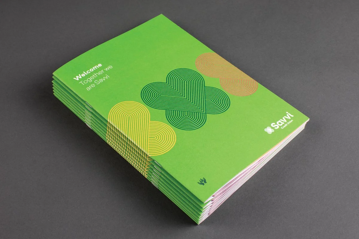 Savvi Credit Union. Green booklet with the title 'Welcome, Together we are Sawi' and a design of colorful line art hearts on the cover, placed on a dark gray surface.
