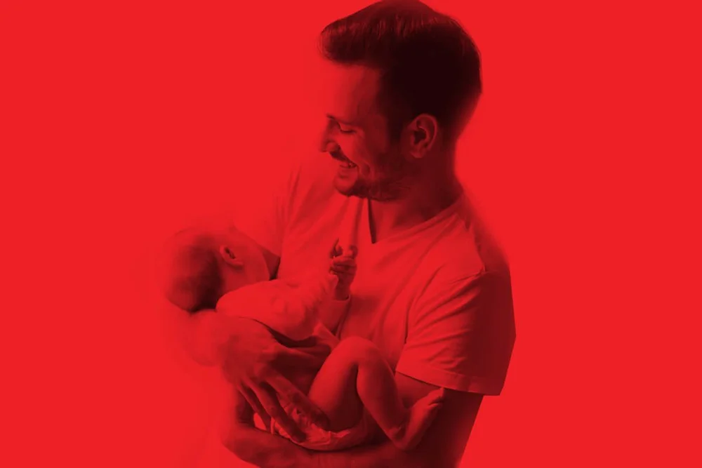 Campaign photograph printed in a single red duotone, a man looking down at the baby in his arms