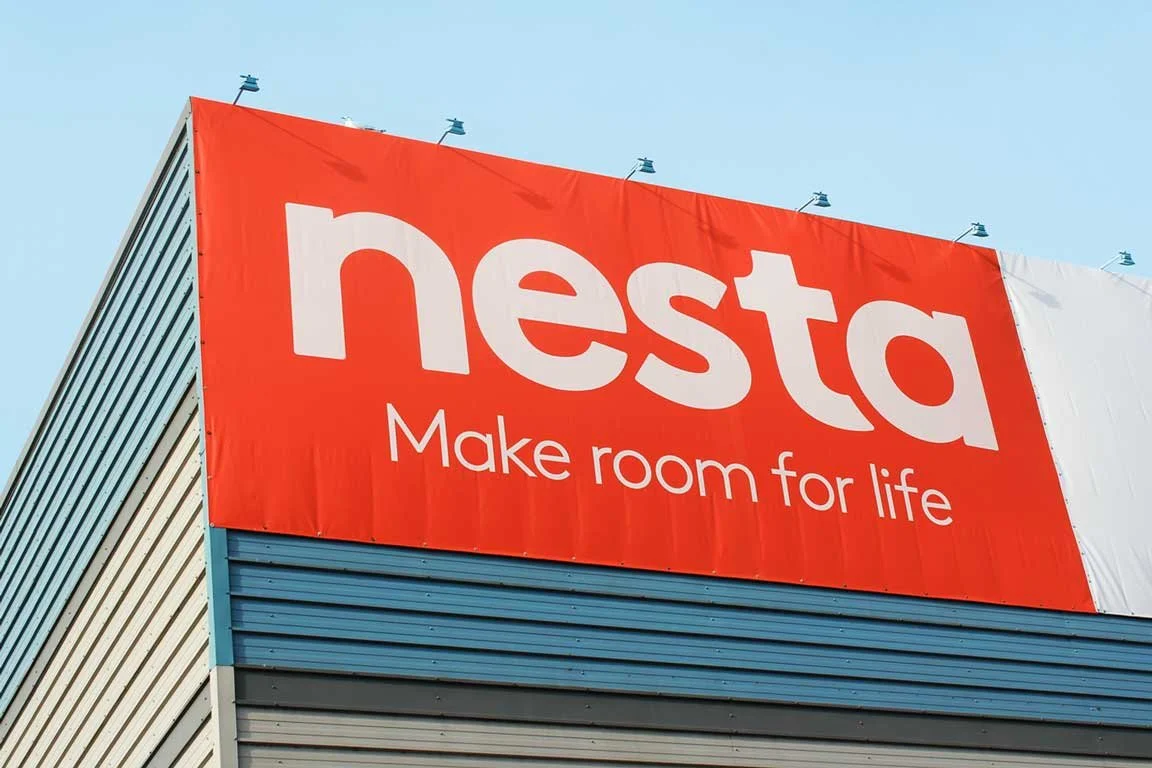 Nesta Storage. Large billboard with a red background displaying the company name 'Nesta' in white letters and the tagline 'Make room for life.' The billboard is mounted on a building with a blue and gray facade under a clear blue sky.