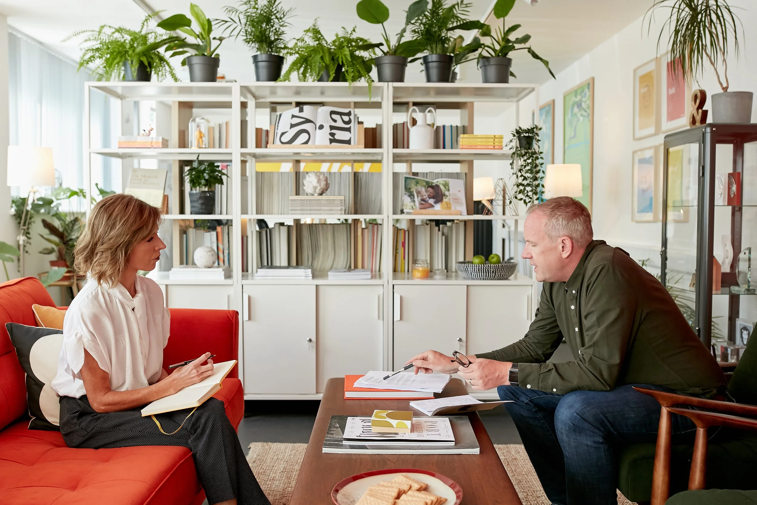 A woman and a man are sitting across from each other in a cozy, well-lit living room. The woman is on an orange sofa, writing in a notebook, while the man is leaning forward, holding glasses and some papers on the coffee table, engaged in conversation. Behind them is a large white bookshelf filled with books and decorative items, with green potted plants on top.
