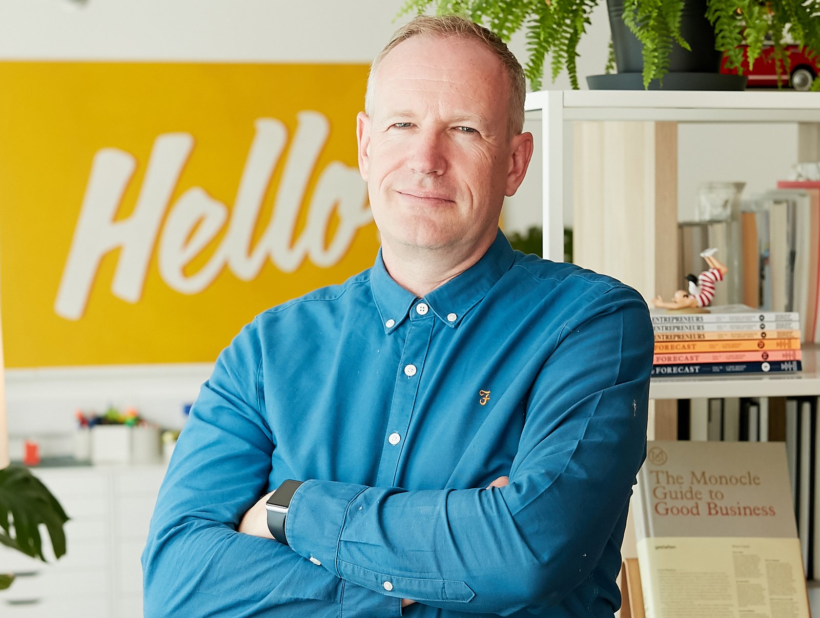 Adam Gallacher. A man with short hair wearing a blue button-up shirt, standing with arms crossed in an office space with a yellow 'Hello' sign on the wall and shelves with books and decorations behind him.