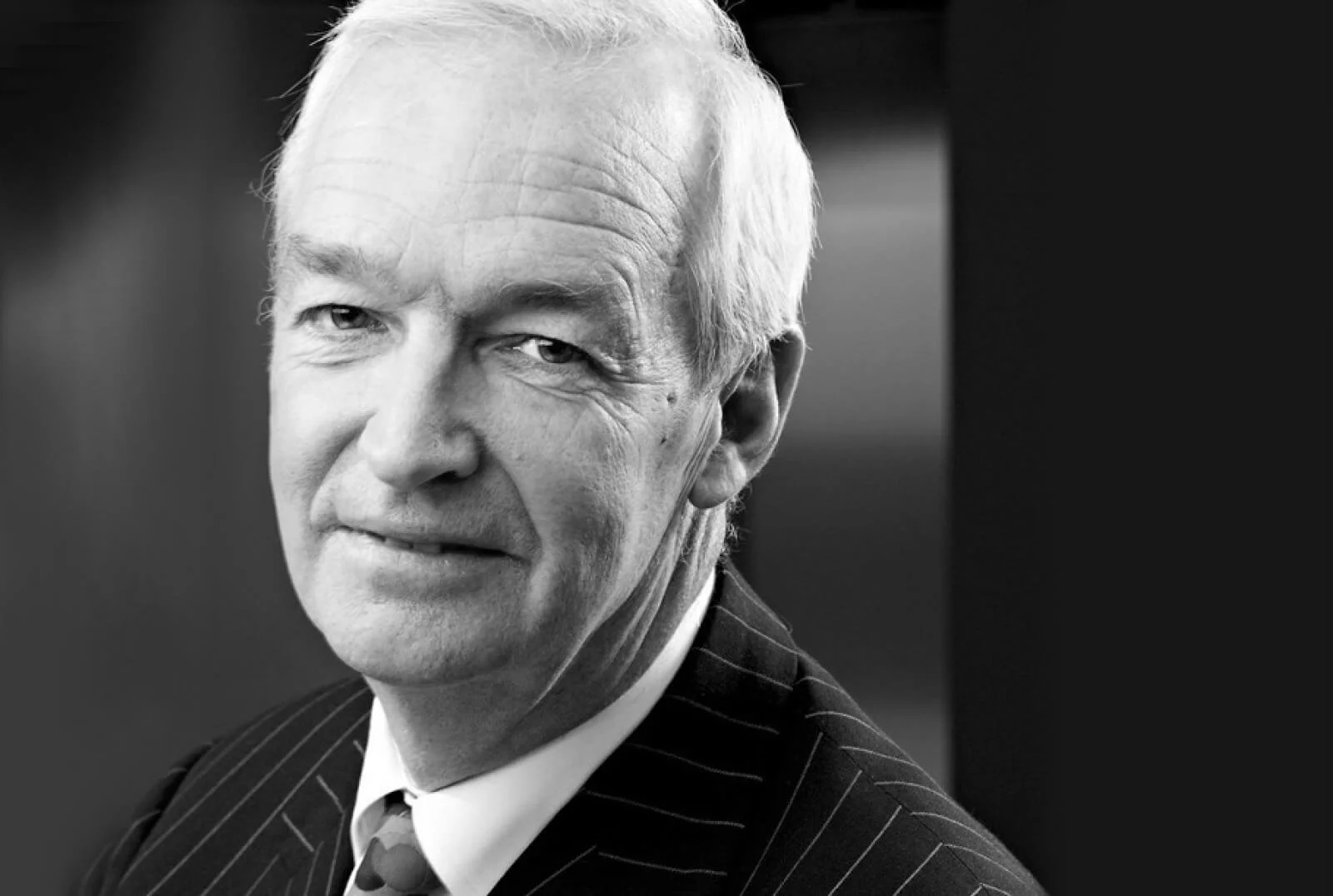 Jon Snow. A black and white portrait photograph of an older man with white hair, wearing a pinstripe suit and a tie, looking slightly to the side with a serious expression.