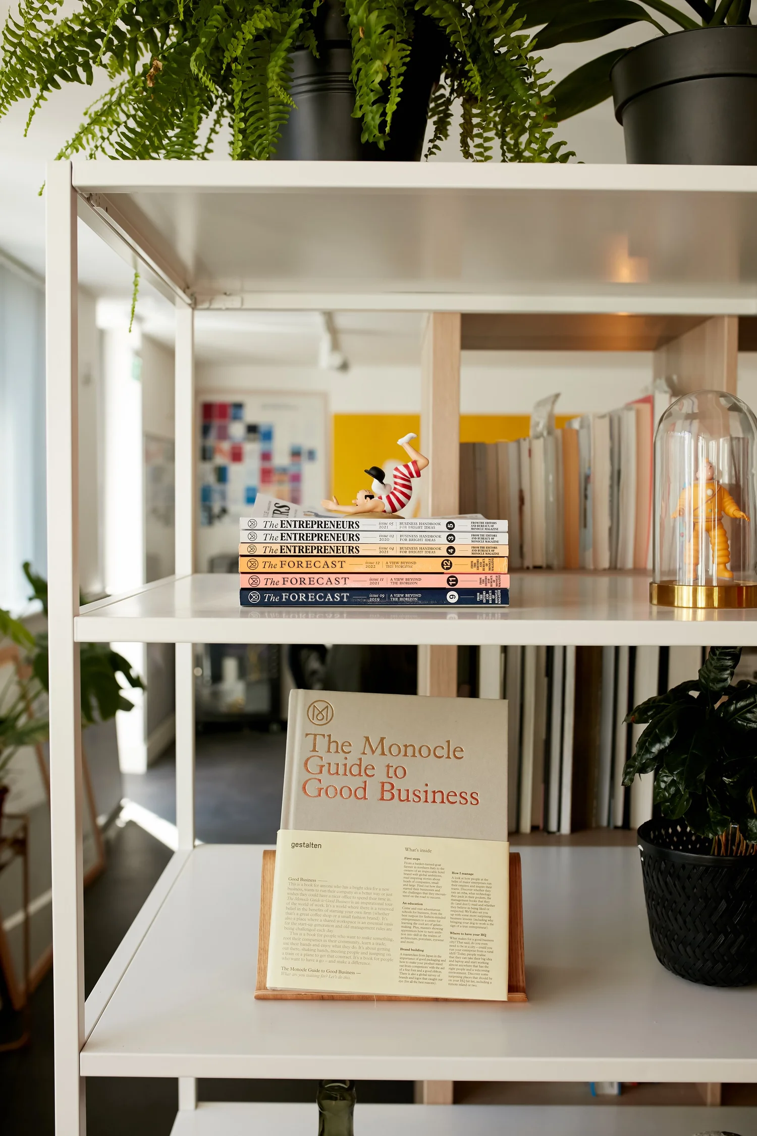 A studio shelf: ferns above, a stack of Monocle Forecast and Entrepreneurs issues, and the Monocle Guide to Good Business open on a stand