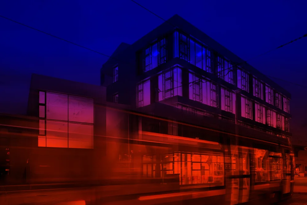 A modern corner building at night with a tram passing in motion blur, graded from deep blue into red