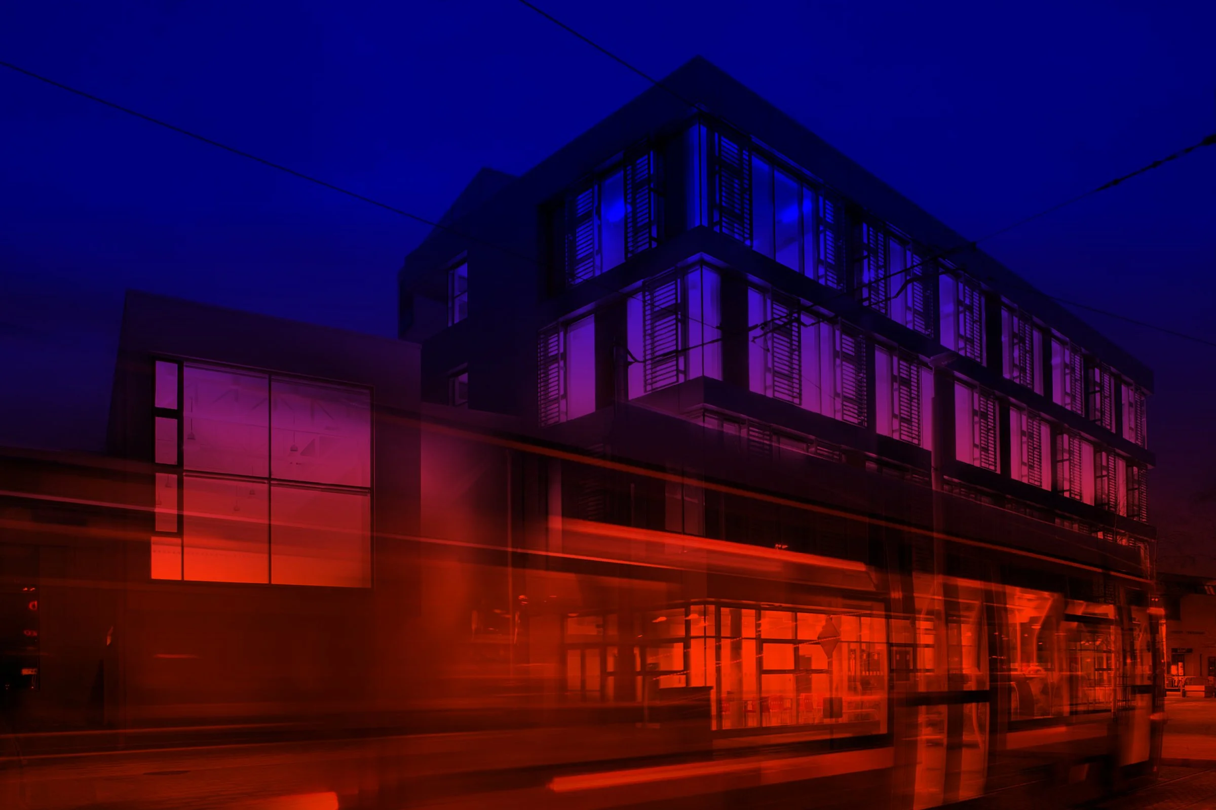 A city corner at night, lit windows above and a tram passing in motion blur below