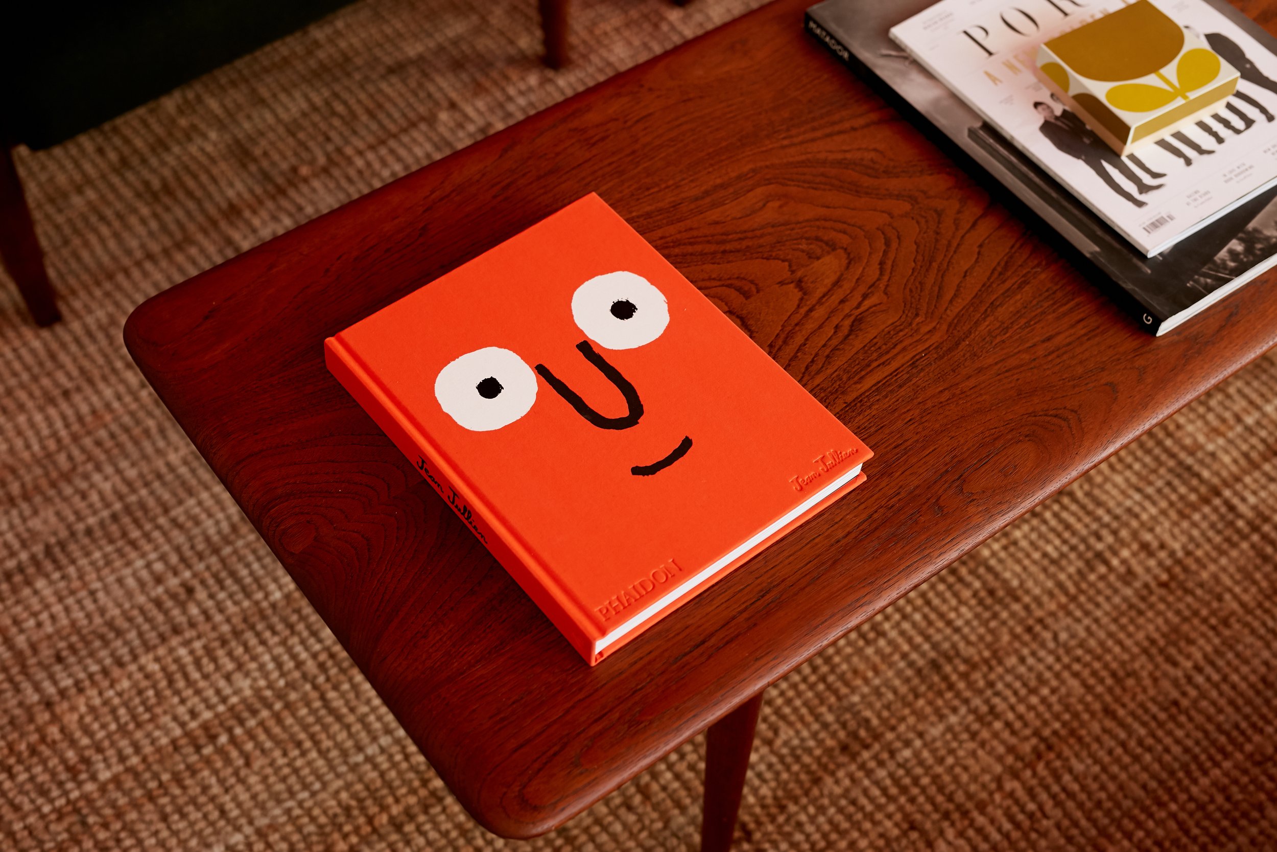 A wooden table with a book featuring a cartoon face with big eyes and a smile, next to a stack of magazines.